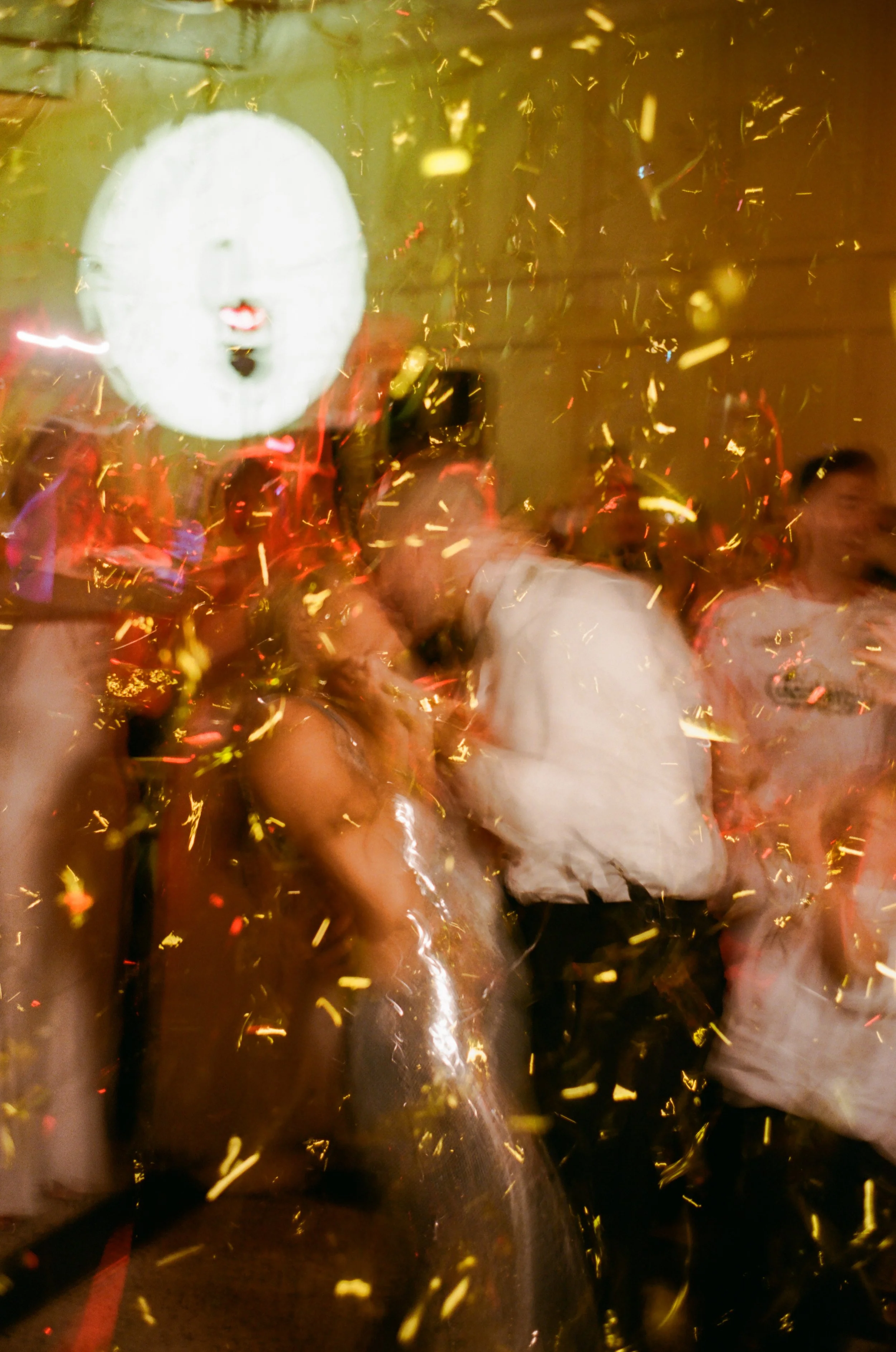 People celebrating at a party with golden confetti and balloons, one person wearing a white shirt, others in casual attire, some holding drinks, and festive lighting in the background.