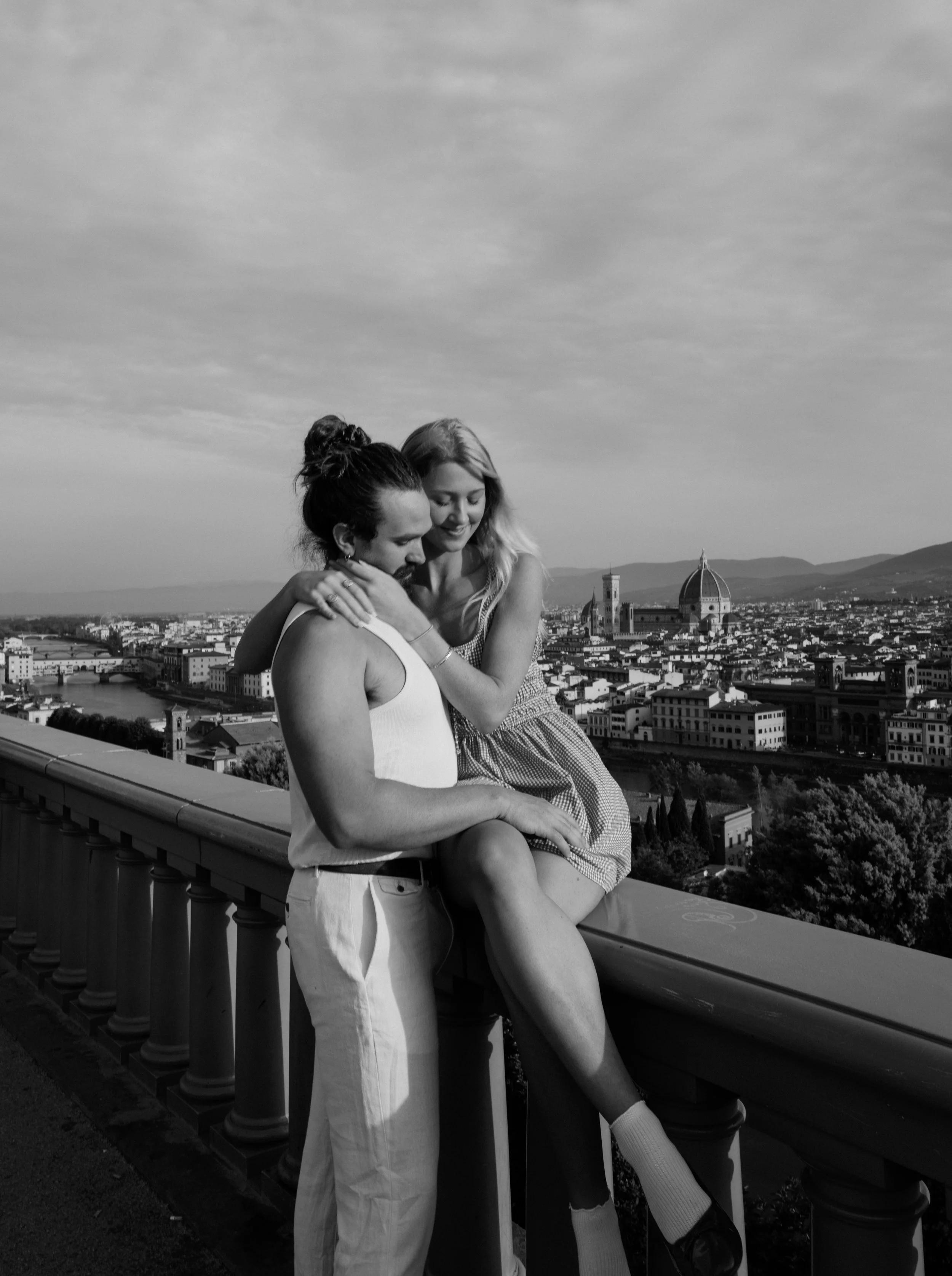 A black-and-white photo of a man and a woman embracing on a bridge or balcony with a scenic cityscape behind them, including a prominent dome and historical buildings, under cloudy skies.