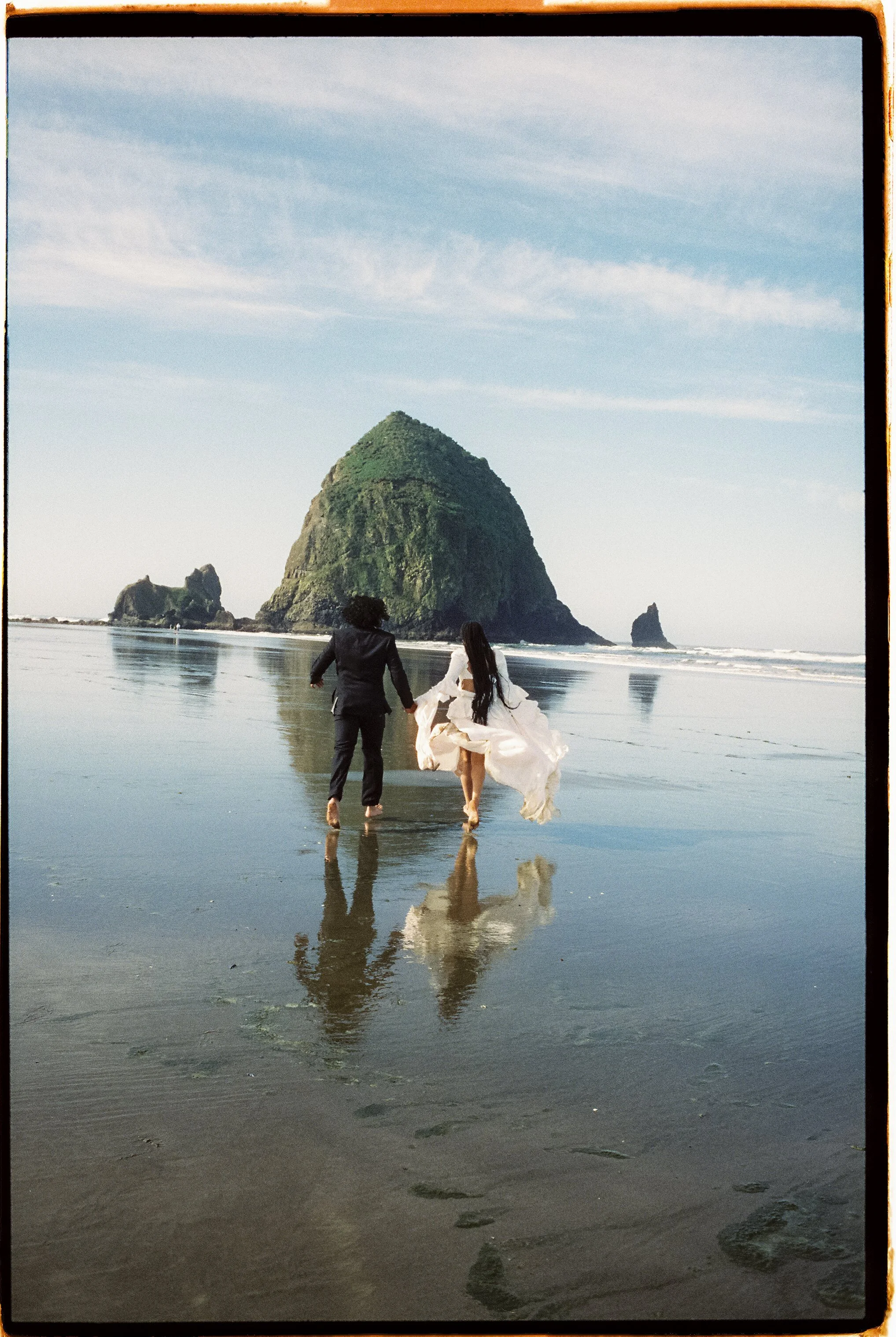 A couple walking hand in hand along the shoreline of a beach with rocks and a large green-covered rock formation in the background, under a partly cloudy sky.