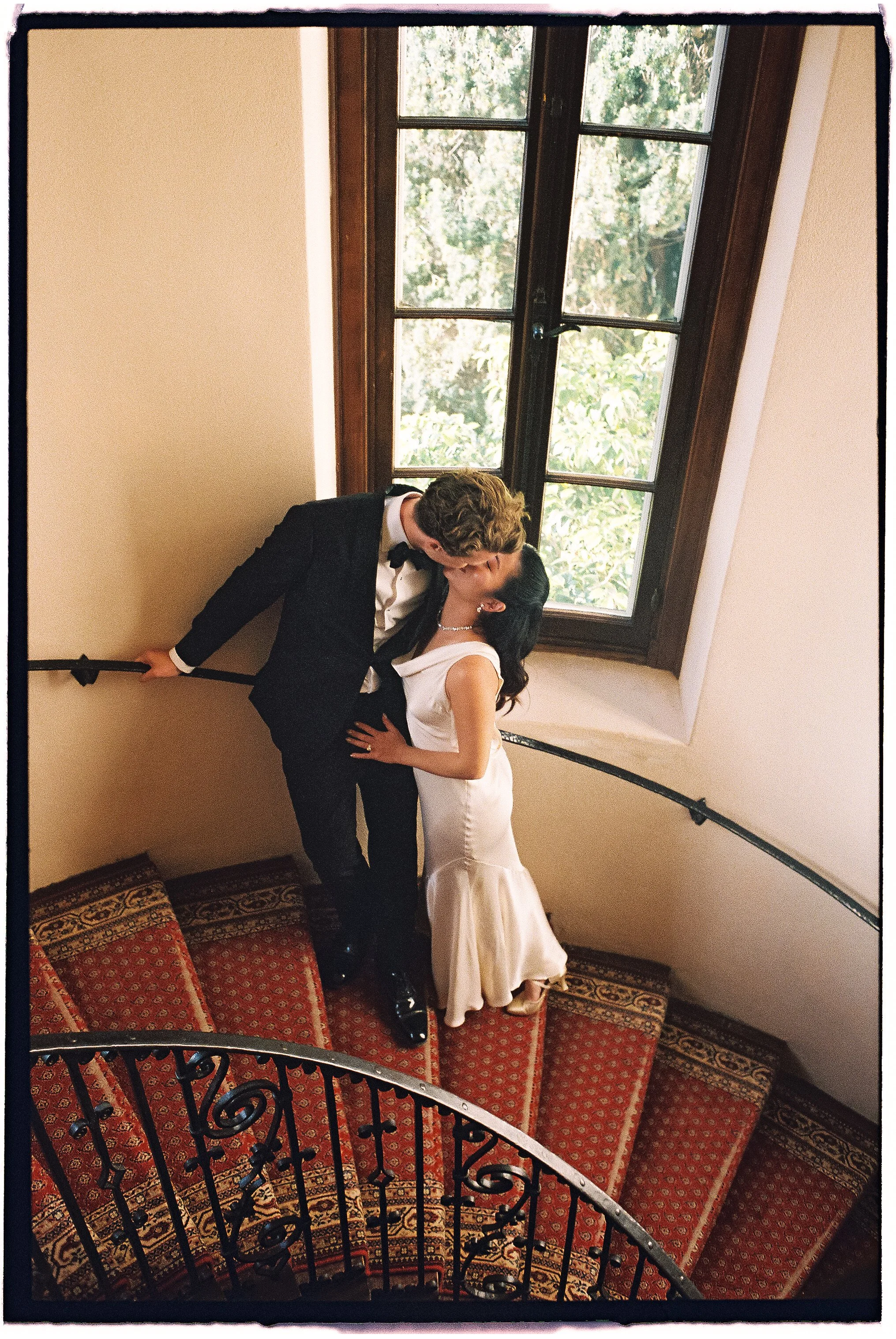 A couple in wedding attire sharing a kiss on a staircase landing near a large window with trees outside. The man is dressed in a tuxedo, and the woman is wearing a white wedding gown. They are standing with the man leaning over the woman, who has her