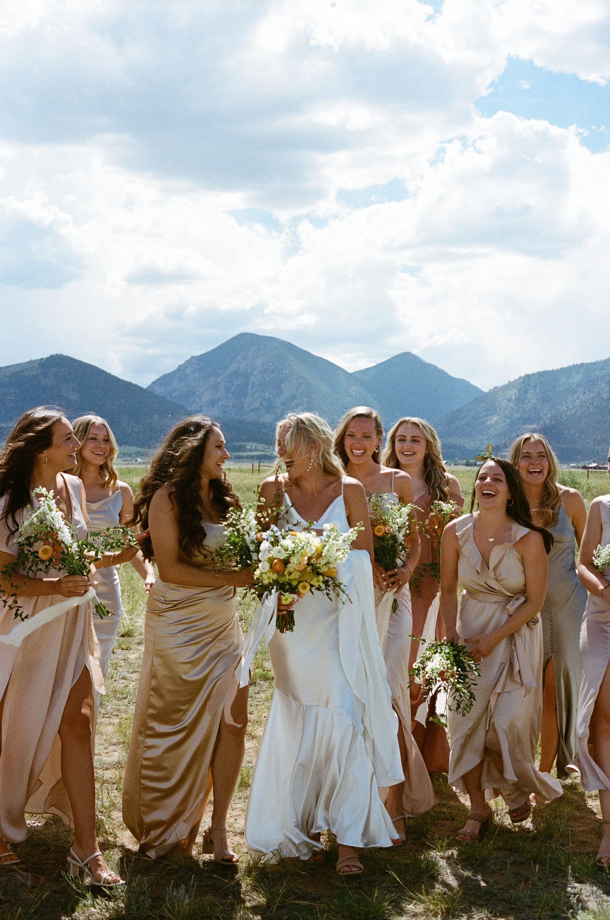 A group of women in formal dresses outdoors, with mountains and cloudy sky in the background, smiling and holding bouquets during a wedding celebration.