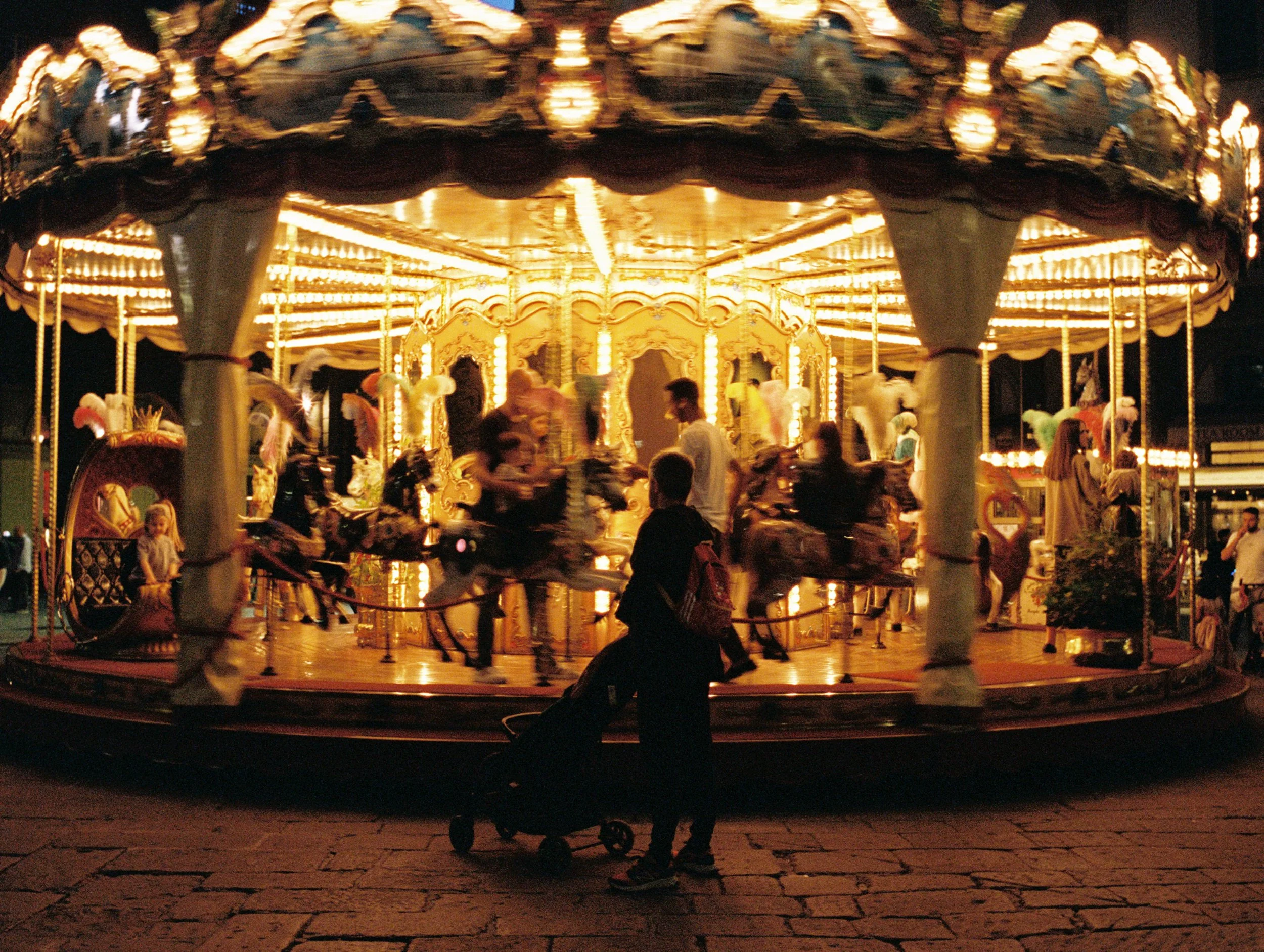 A brightly lit vintage carousel at night with ornate decorations and children riding the horses, and a young boy standing nearby with a stroller.