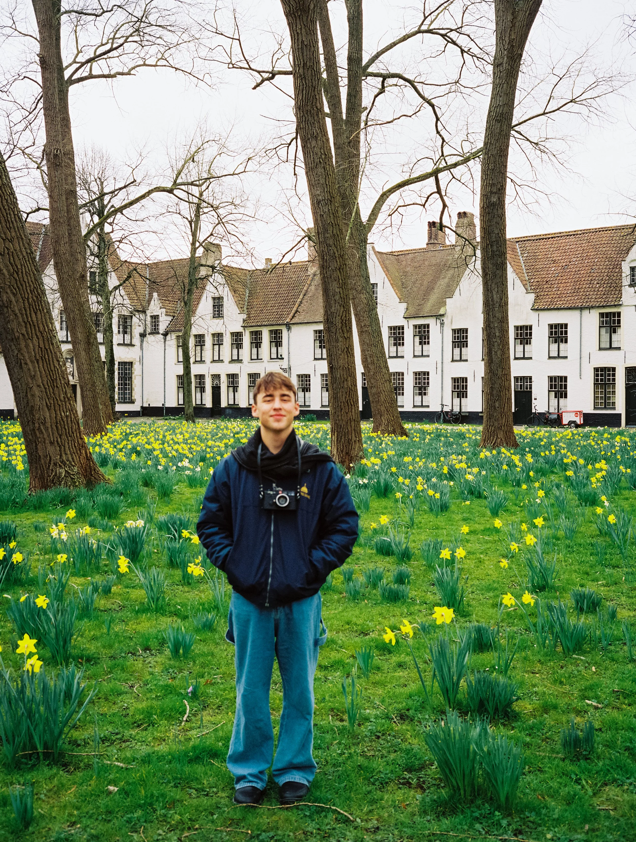 A young man with curly hair wearing a dark jacket and jeans standing outdoors on grass, surrounded by yellow flowers and tall leafless trees, with white buildings in the background.