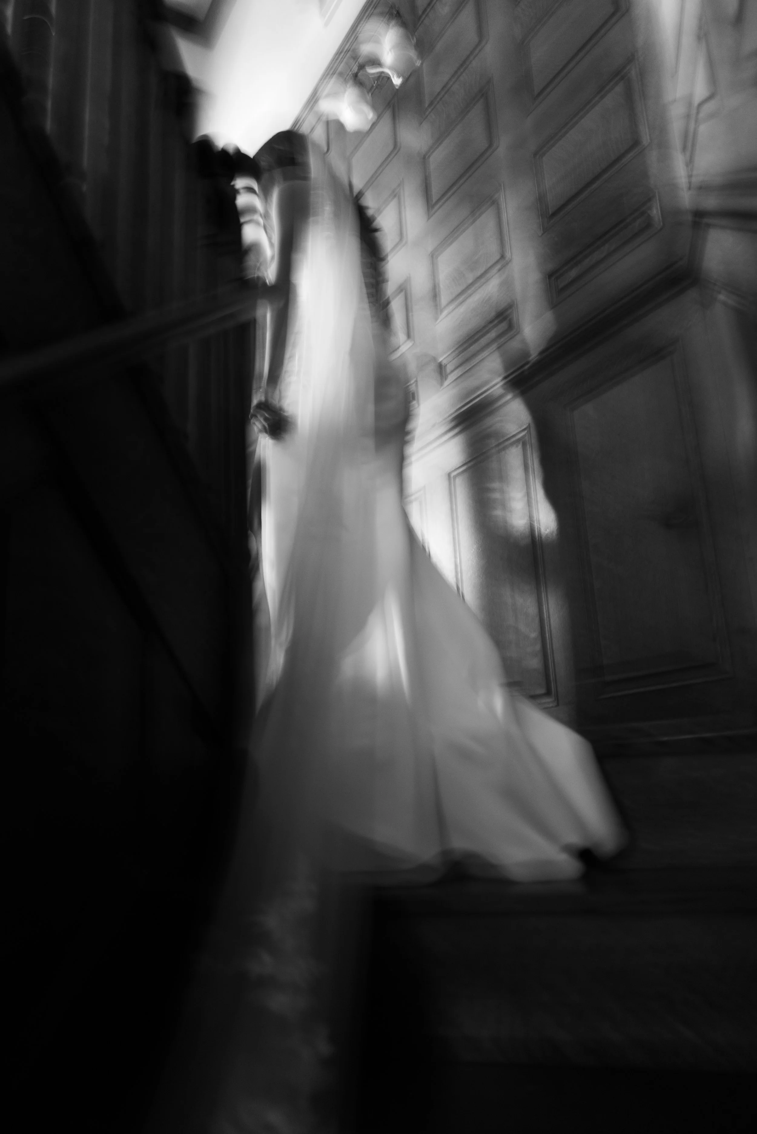 A black and white photo of a woman in a wedding dress ascending a staircase with ornate wooden paneling on the wall.