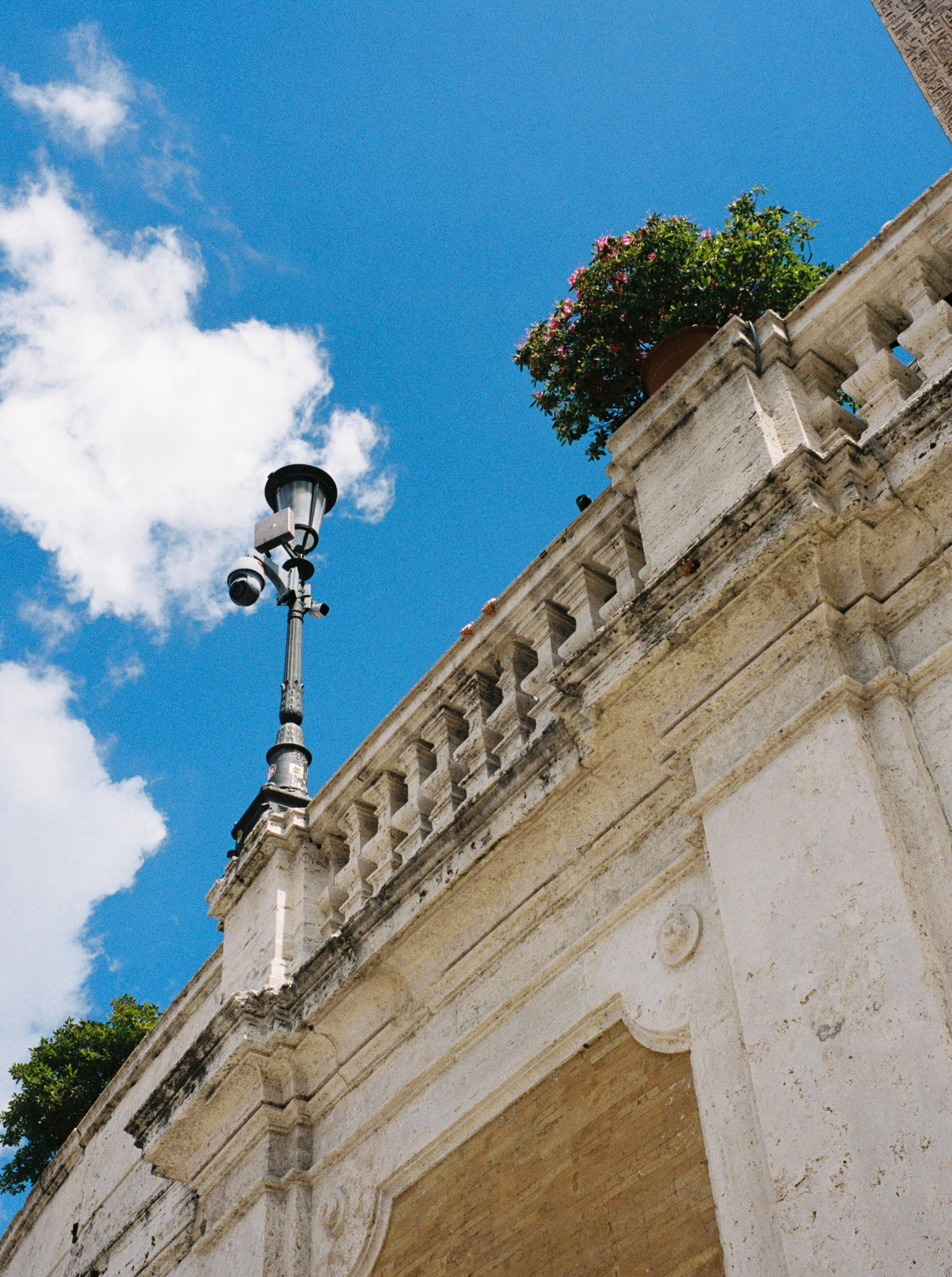 A historic building with a decorative stone balustrade, a potted plant with pink flowers, a streetlight with security cameras, and a bright blue sky with some clouds.