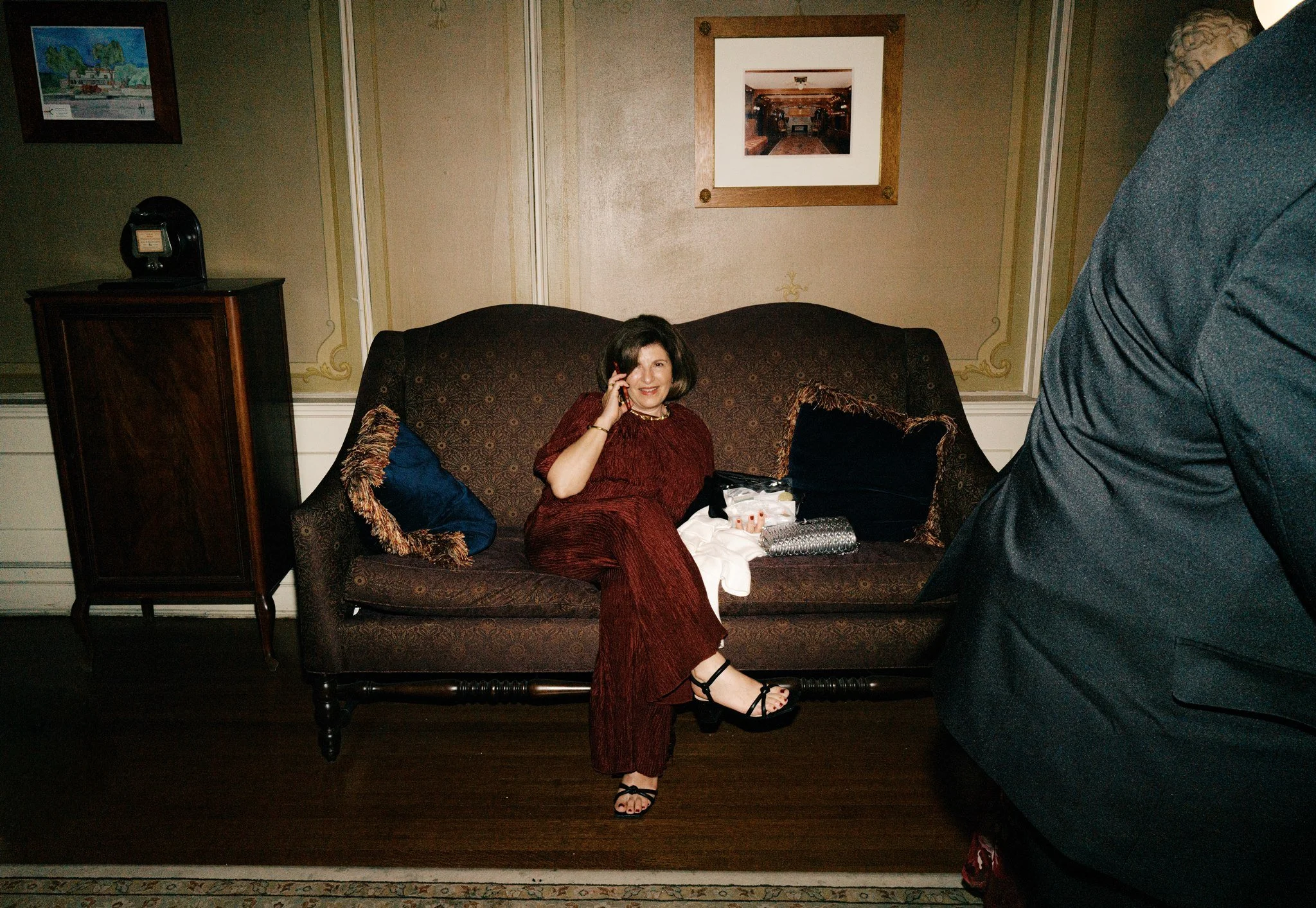 A woman with dark hair wearing a red dress and black sandals, sitting on a patterned sofa, smiling, talking on a phone, with a woman in a black jacket partially visible in the foreground.