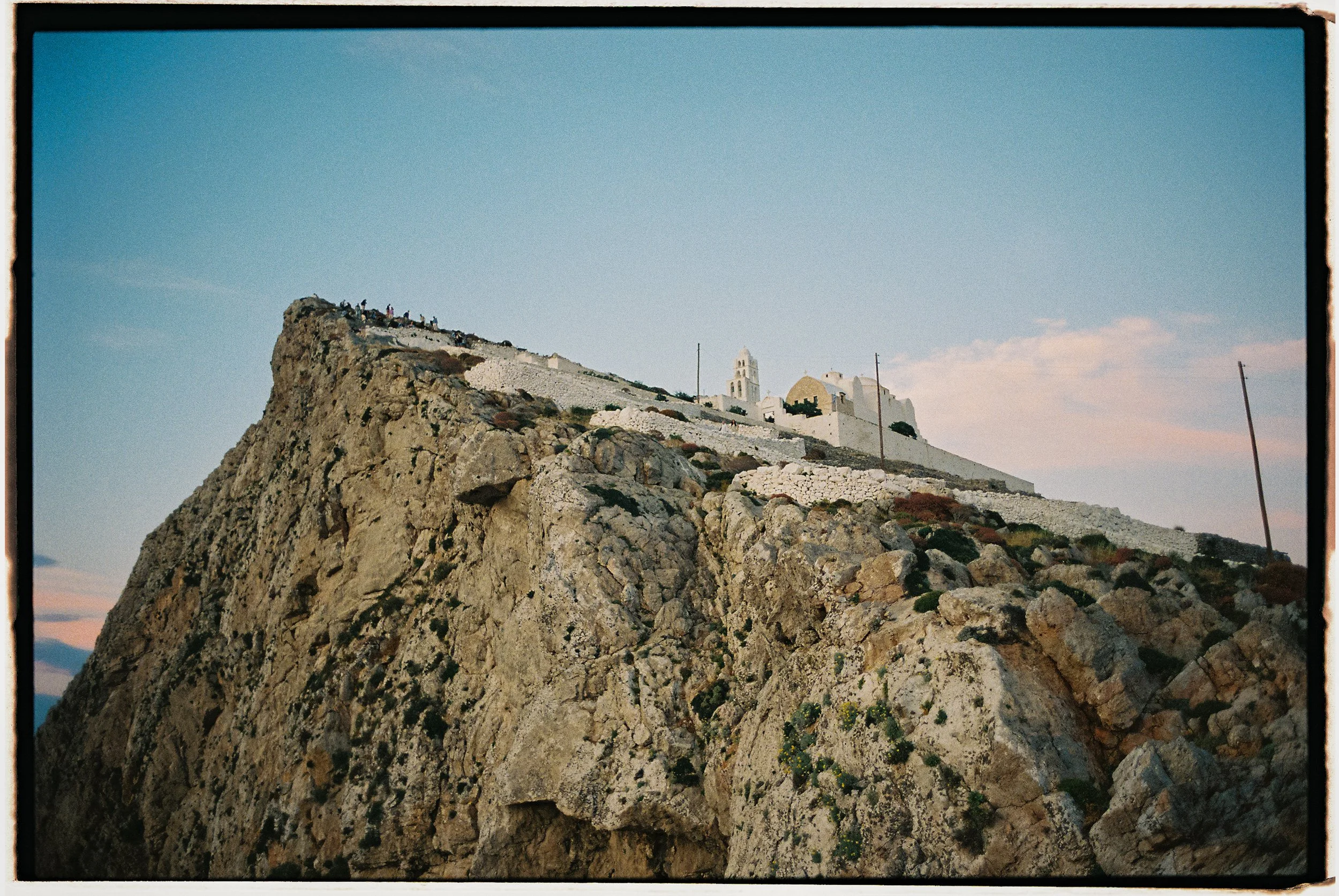 A rocky hill with a white church, bell tower, and buildings atop. The sky is clear and blue.
