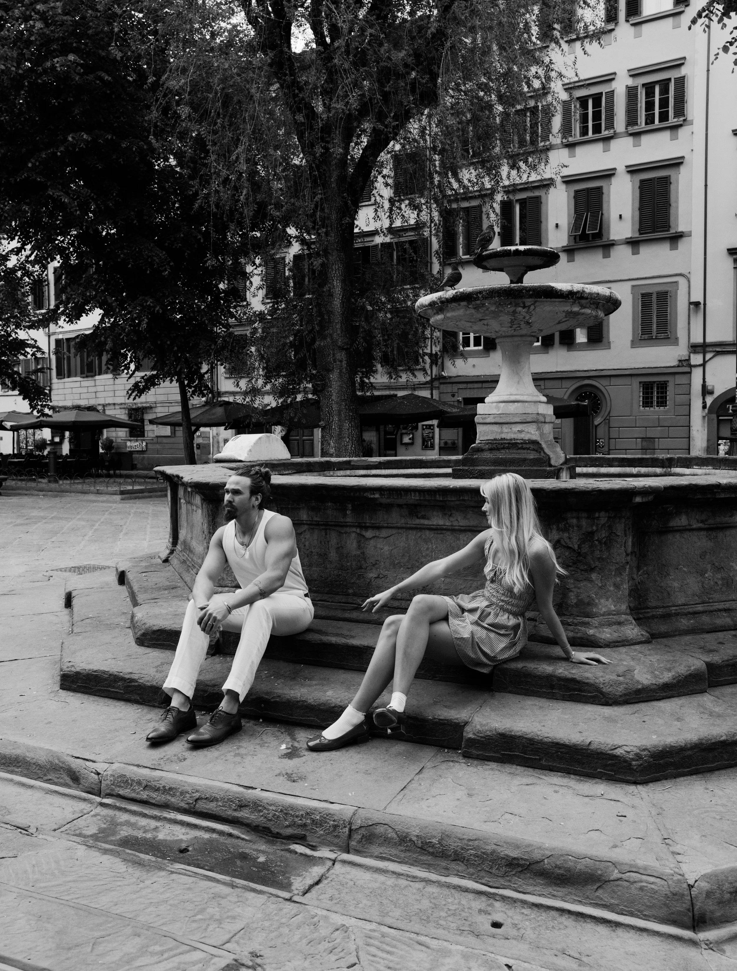Two people sitting on an old stone fountain in an urban square, with a large tree and a multi-story building with shuttered windows in the background. The man has dark hair, a beard, and is wearing light-colored pants and a sleeveless top. The woman 