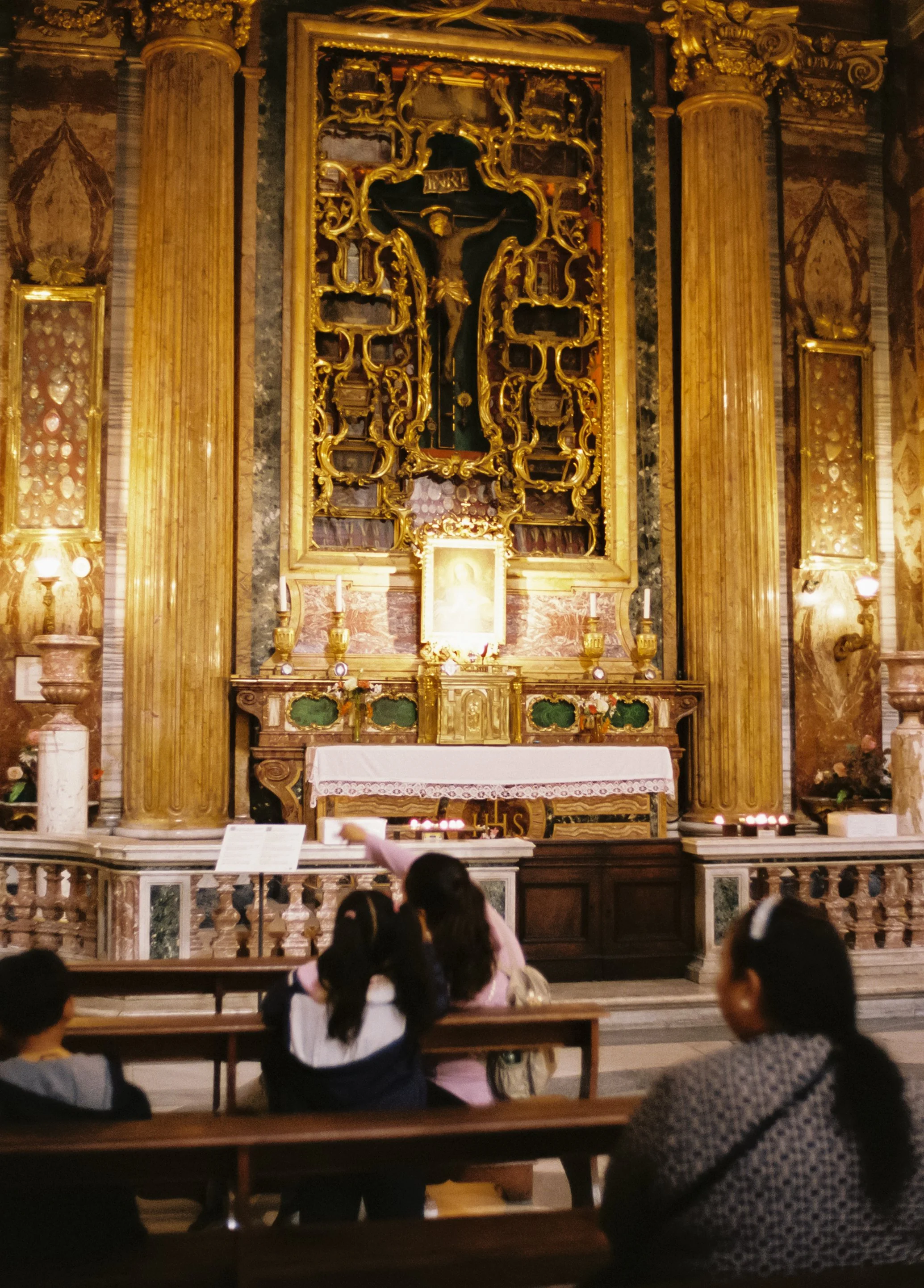 Inside a church, people seated on benches facing an ornate altar with a crucifix and religious icons.