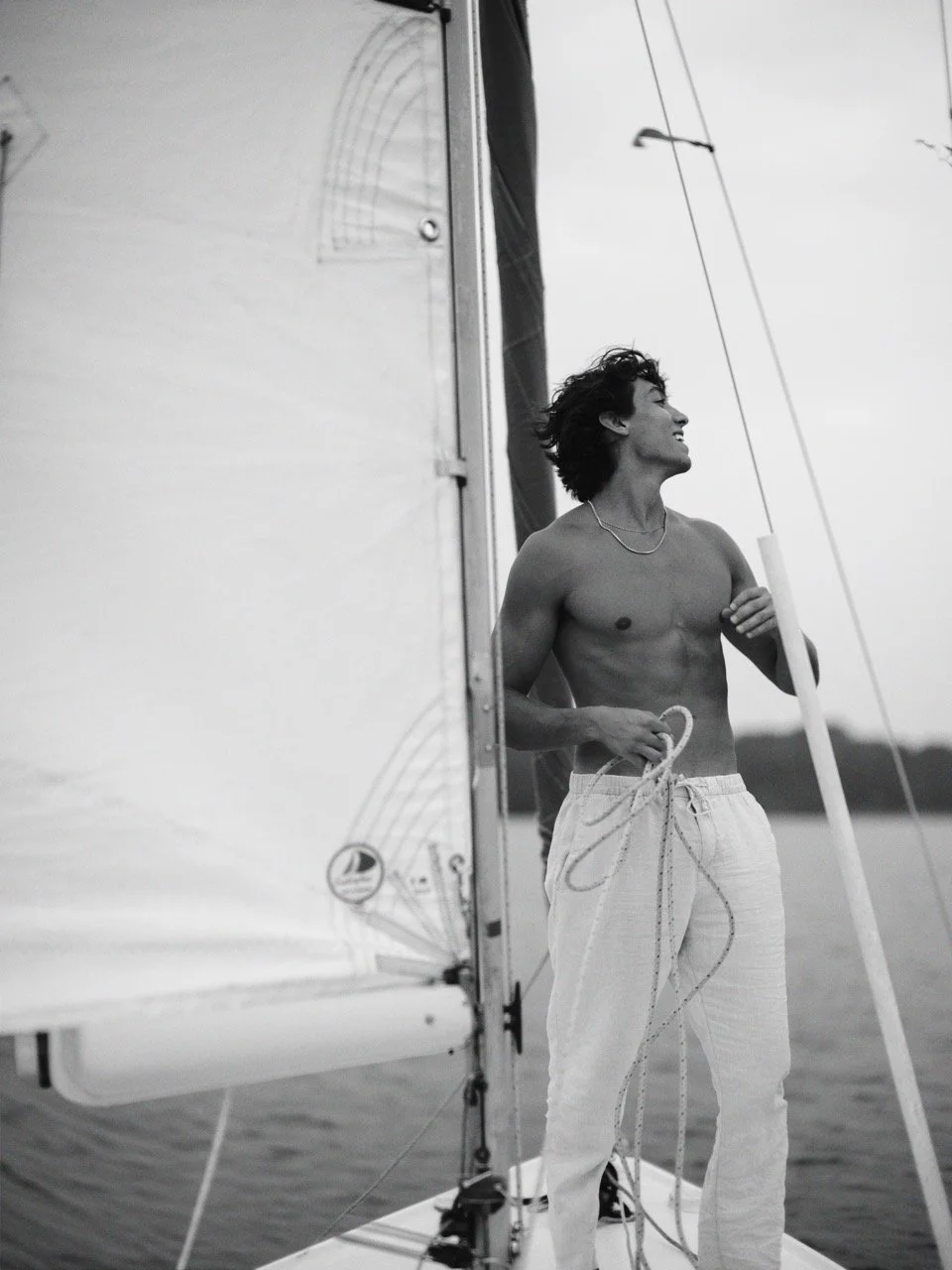 A shirtless young man with dark, curly hair smiling, standing on a sailboat holding ropes, with water in the background.