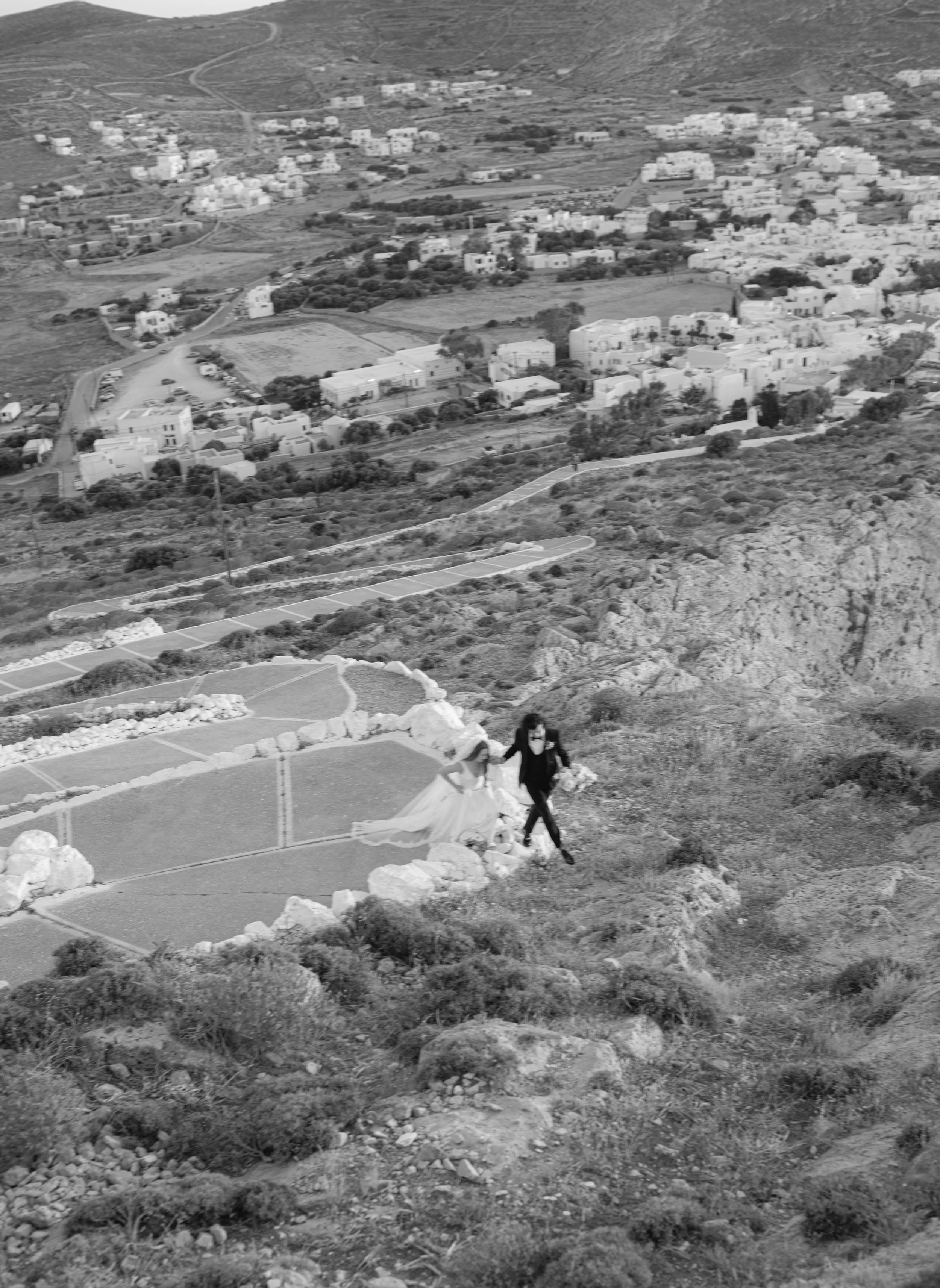 A person walking on a rocky hilltop overlooking a valley with a small village of white buildings, some greenery, and winding roads.