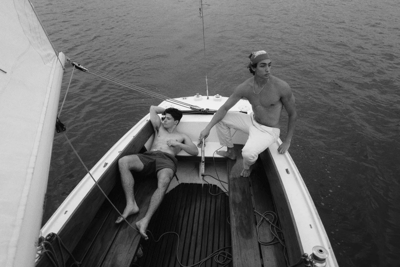 Two young men on a small sailboat; one is lying down and the other is sitting, both shirtless, on a body of water.