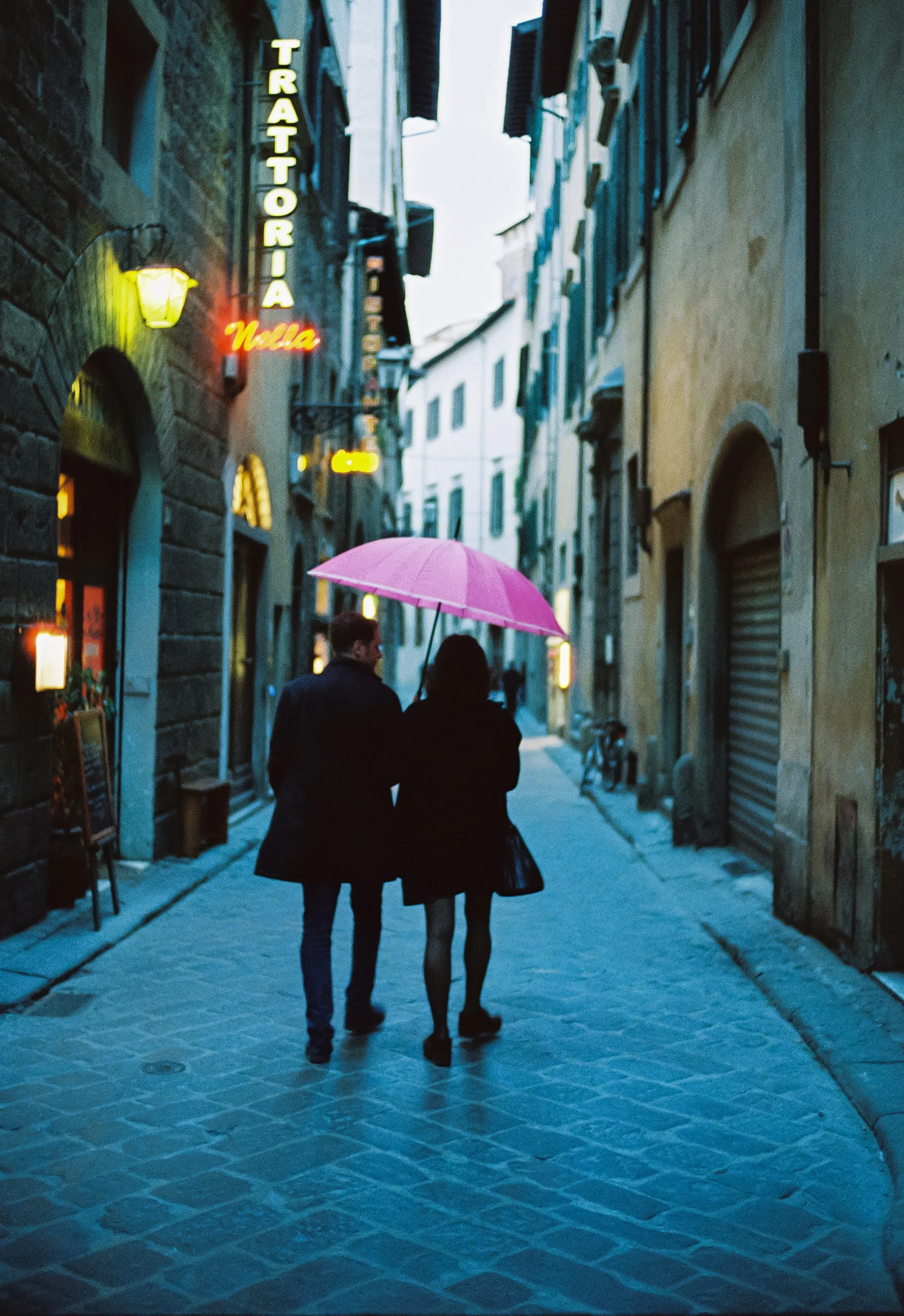 Couple walking together on a cobblestone street at dusk, sharing a pink umbrella, surrounded by European-style buildings with neon signs.