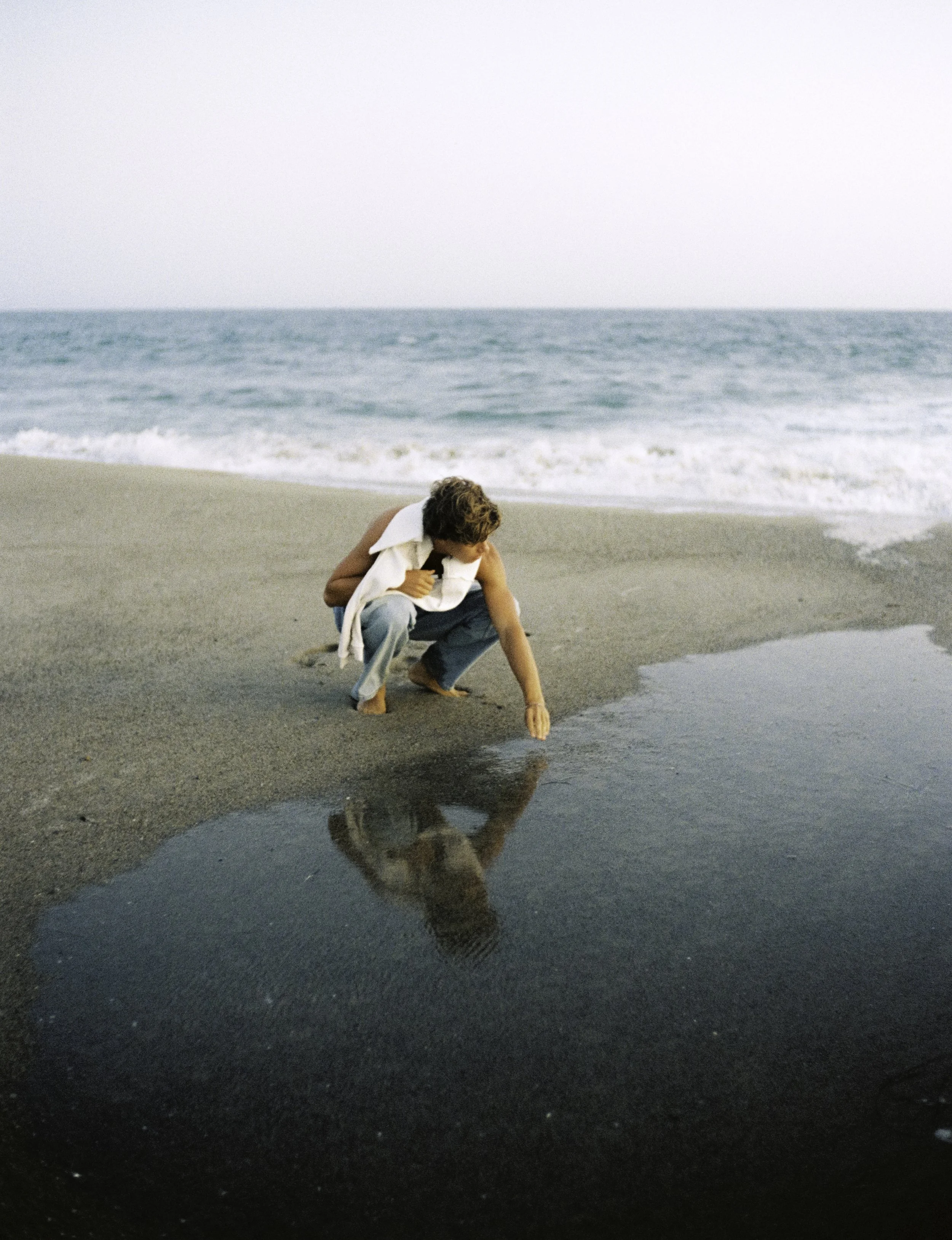 A man crouched down on a sandy beach near the water's edge, touching the small pool of water in front of him, with the ocean and sky in the background.