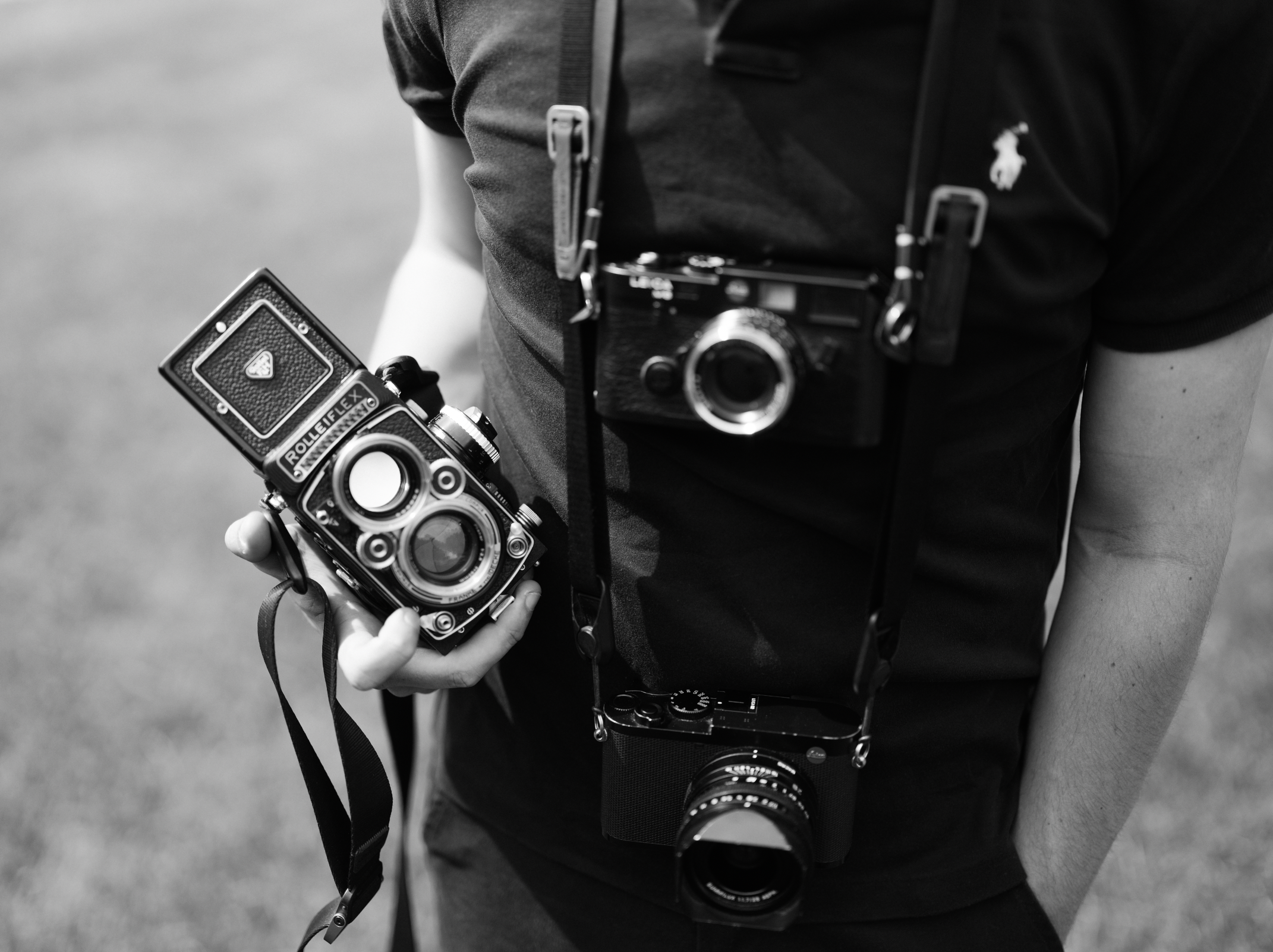 Person holding a vintage camera in one hand while wearing another camera around their neck, dressed in a dark shirt with a small Polo Ralph Lauren logo.