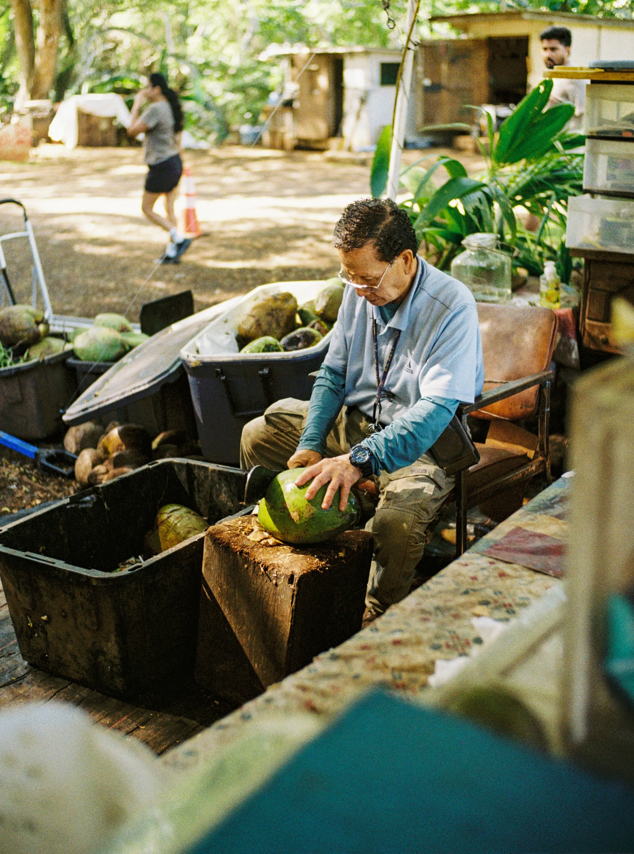 A man peeling a watermelon outdoors, sitting on a wooden chair with a log in front of him. There are crates of watermelons around him and a woman in the background walking past, with greenery and small structures behind her.