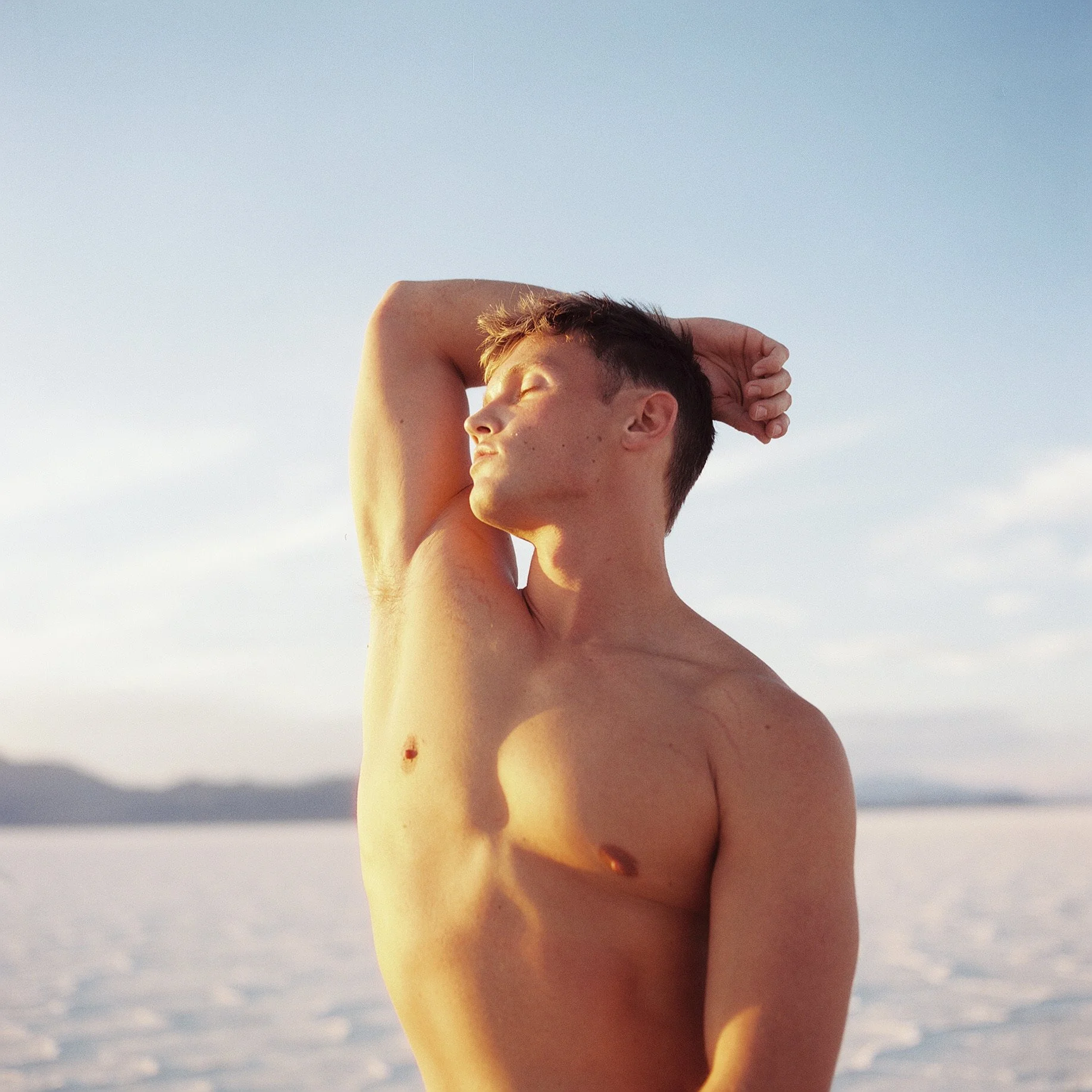 A young shirtless man with short dark hair standing outdoors with a vast white landscape and a mountain range in the background, eyes closed and one arm raised behind his head, under a clear sky.