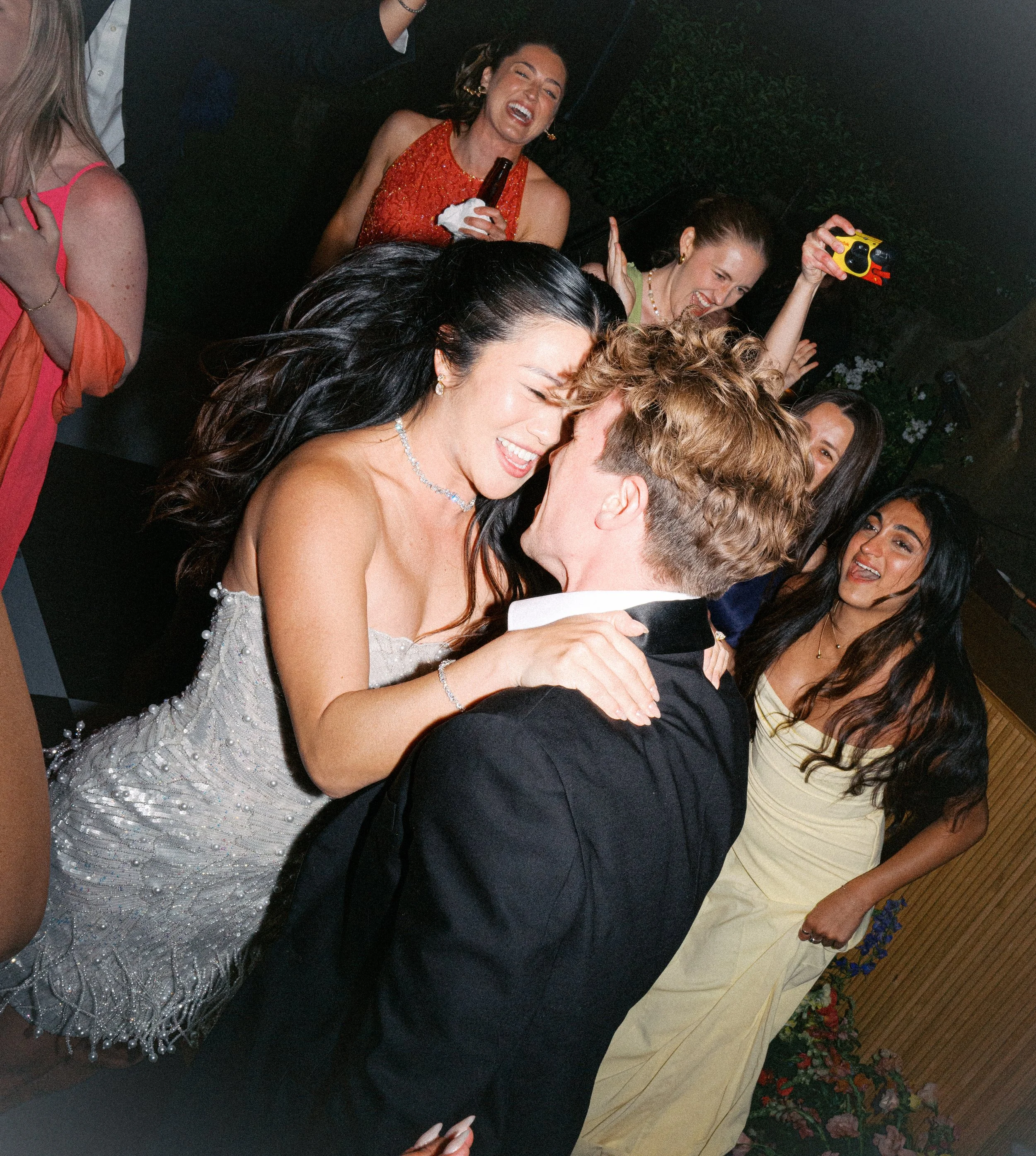 A group of people at a celebration, with a woman in a white dress and diamond necklace and man in formal attire sharing a kiss, surrounded by women taking photos and smiling.