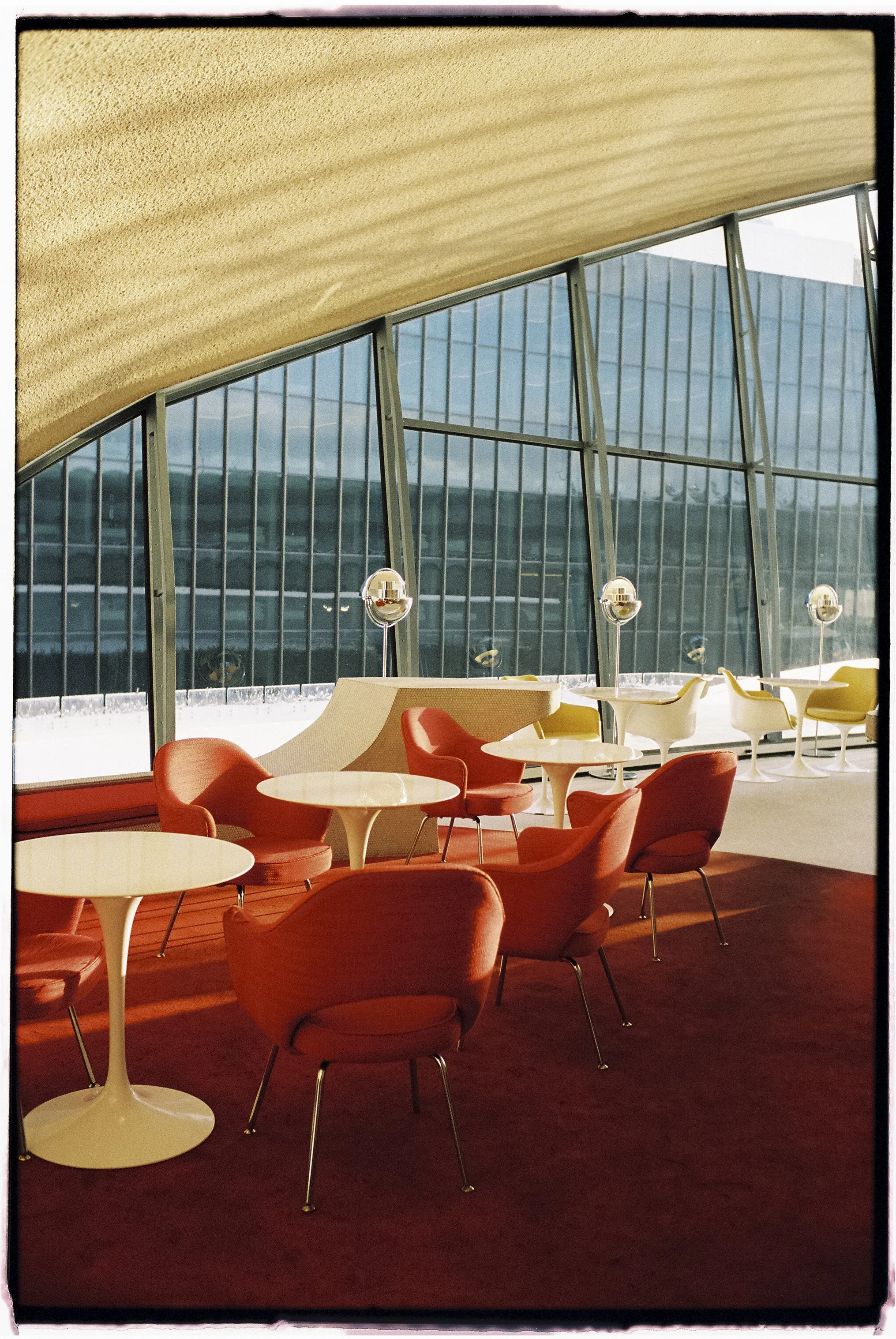 Interior of an airport lounge with red armchairs and white round tables, large windows with a view of modern glass buildings, and curved ceiling design.