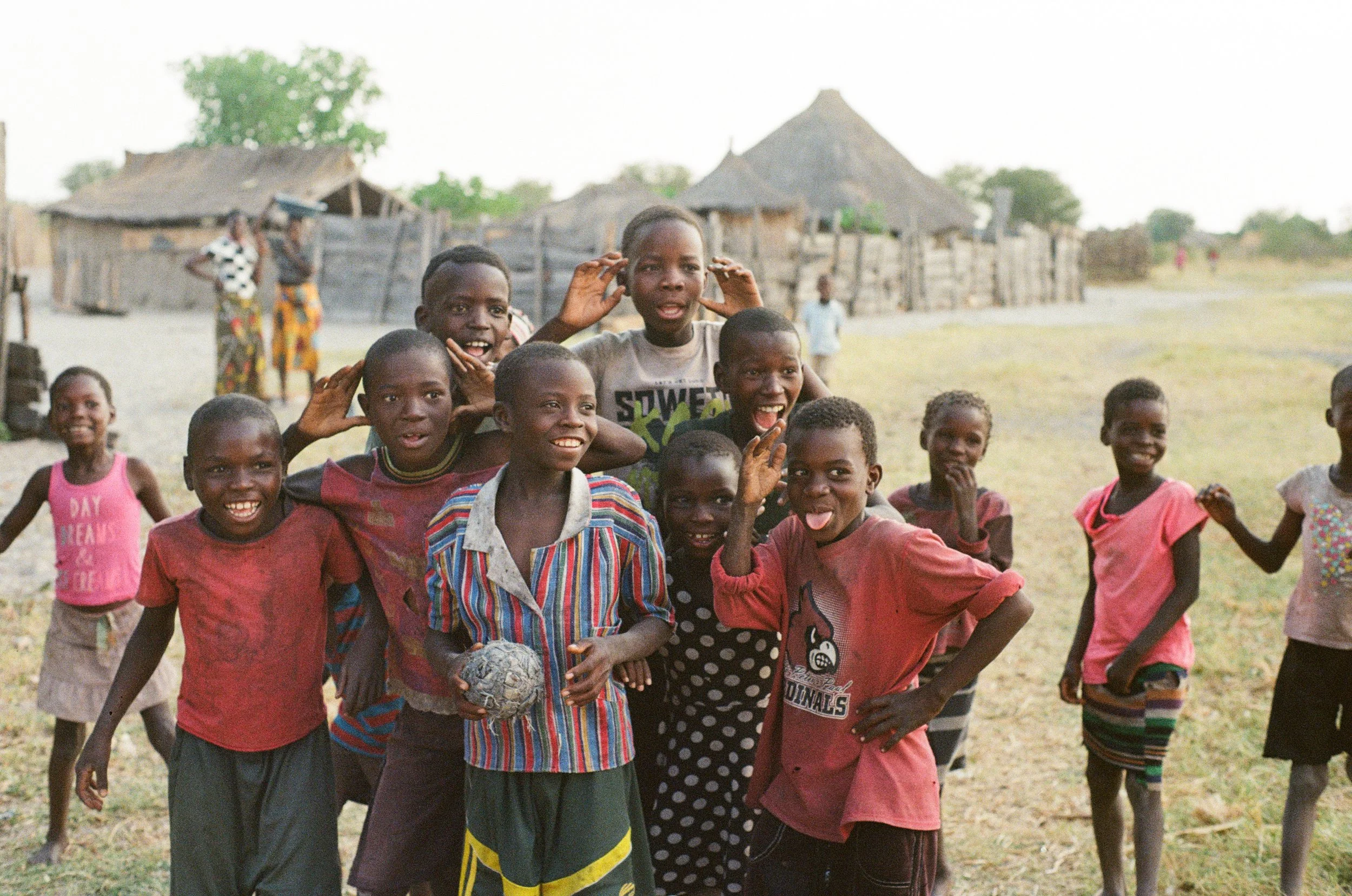 A group of children playing and smiling outdoors in a rural village with traditional thatched huts in the background.