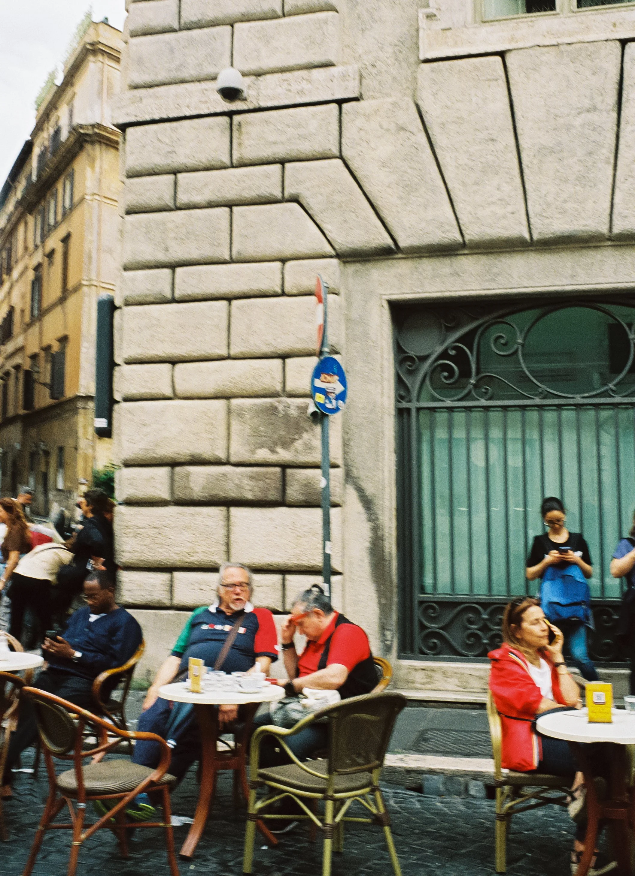 People sitting and standing outside at a cafe on a city street, next to a large stone building with ornate window grille, some using their phones, with street signs visible on the corner.