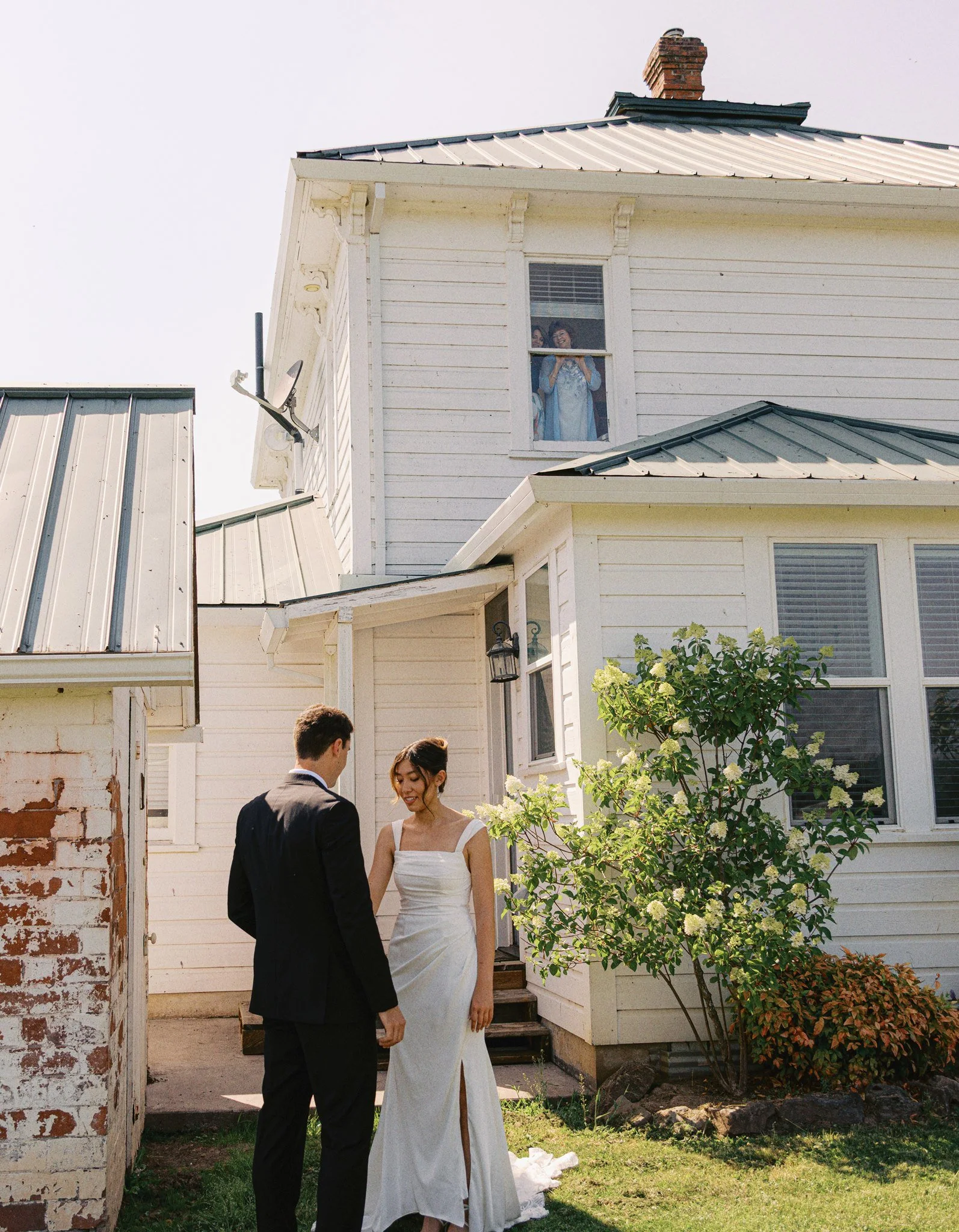 A bride and groom are standing outside a white house with a metal roof, holding hands and looking at each other. A woman is visible through a window smiling, while sunlight illuminates the scene, and lush green plants are in the foreground.