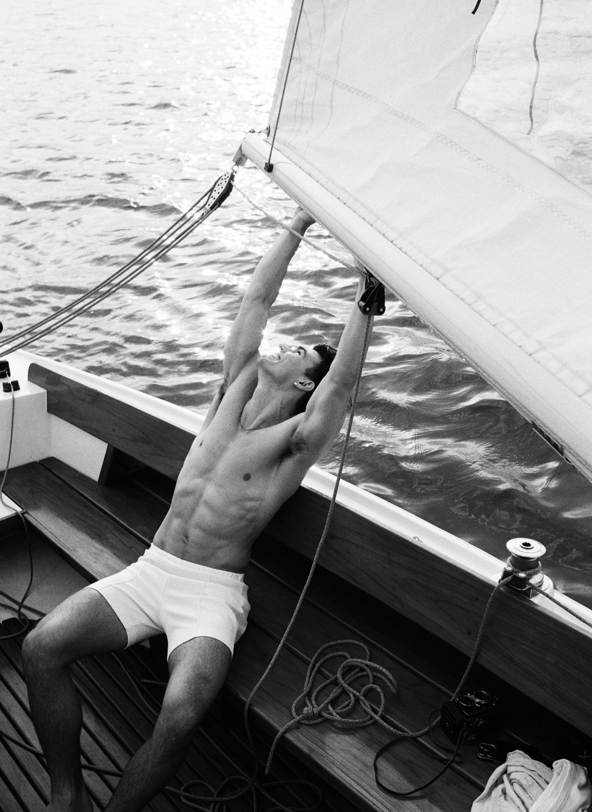 A shirtless man with short dark hair and wearing shorts holding onto a sailing boat's sail, smiling as he adjusts or secures it while sitting on a boat with water in the background.