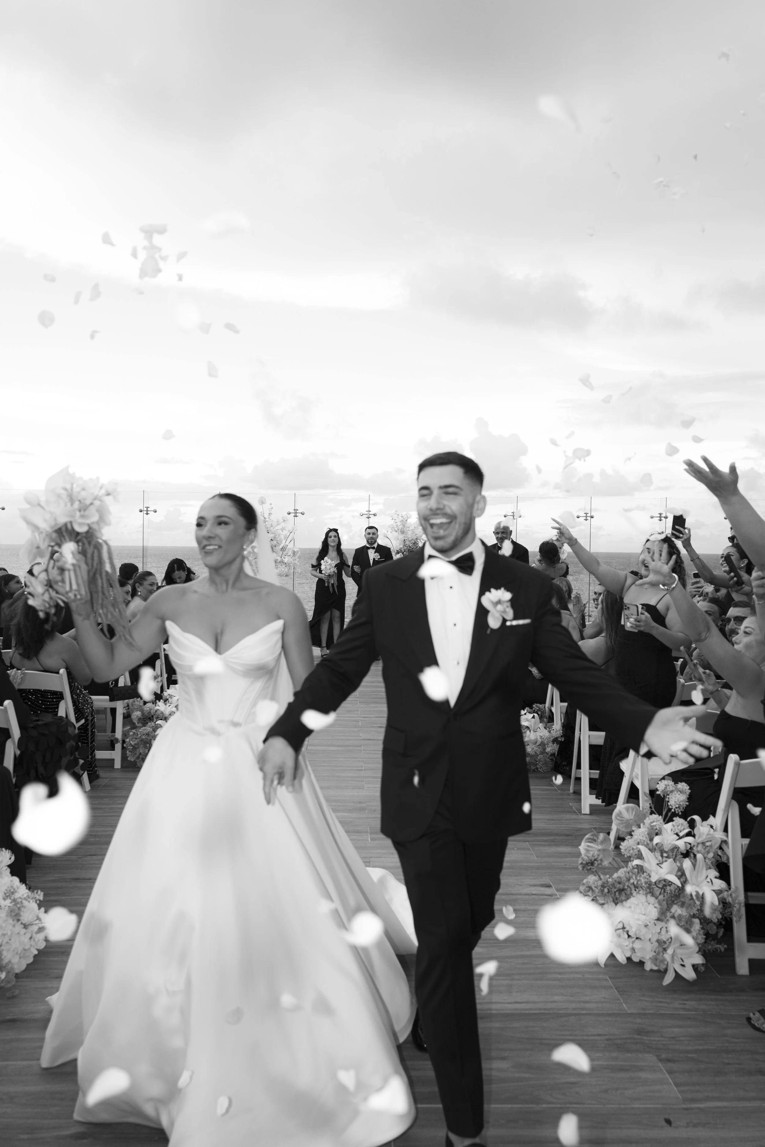 A bride and groom celebrating at their wedding, with guests throwing flower petals, outdoor setting, cloudy sky, and ocean view in the background.