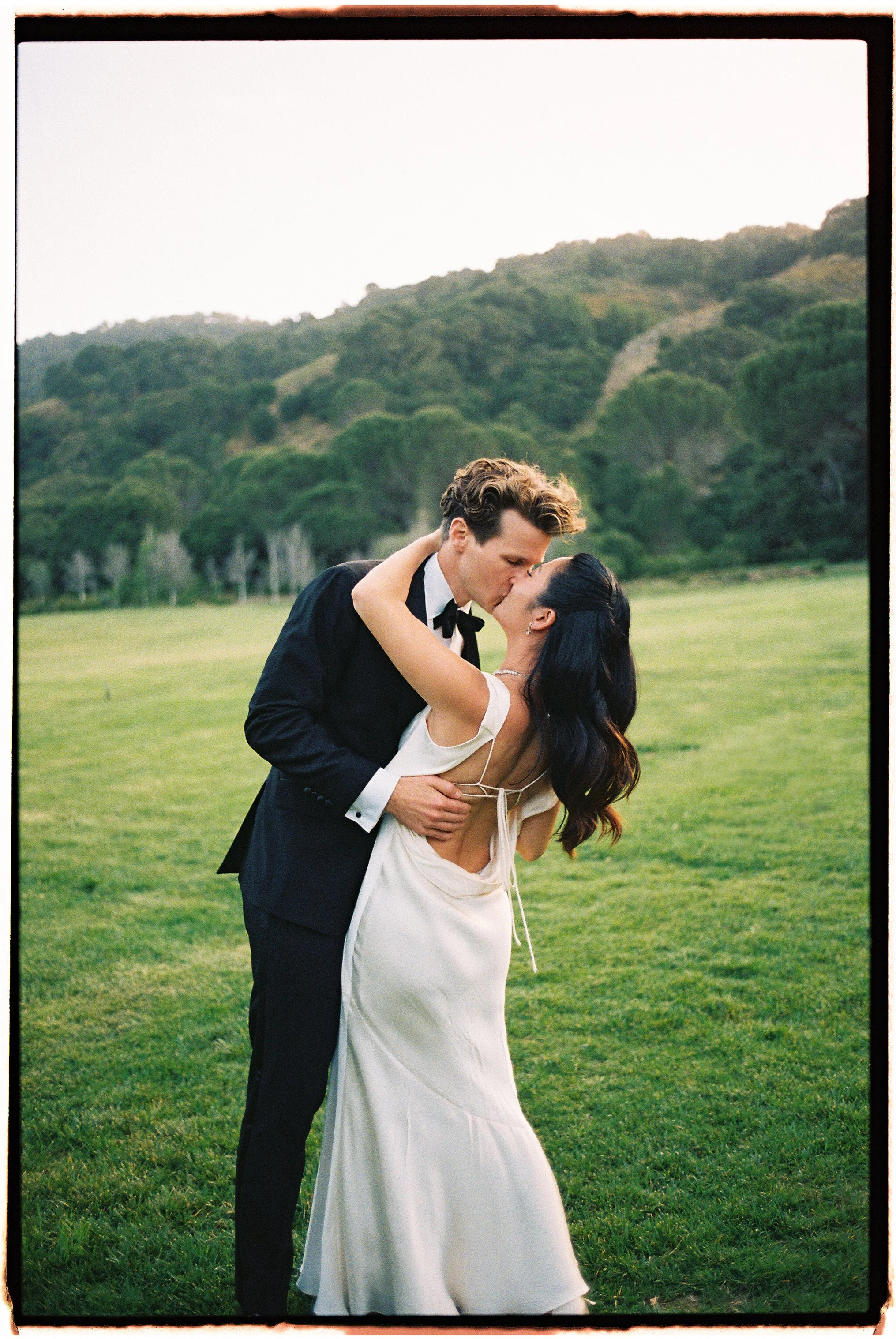 A bride and groom sharing a kiss in an outdoor lush grassy field with green hill and trees in the background, during daylight.