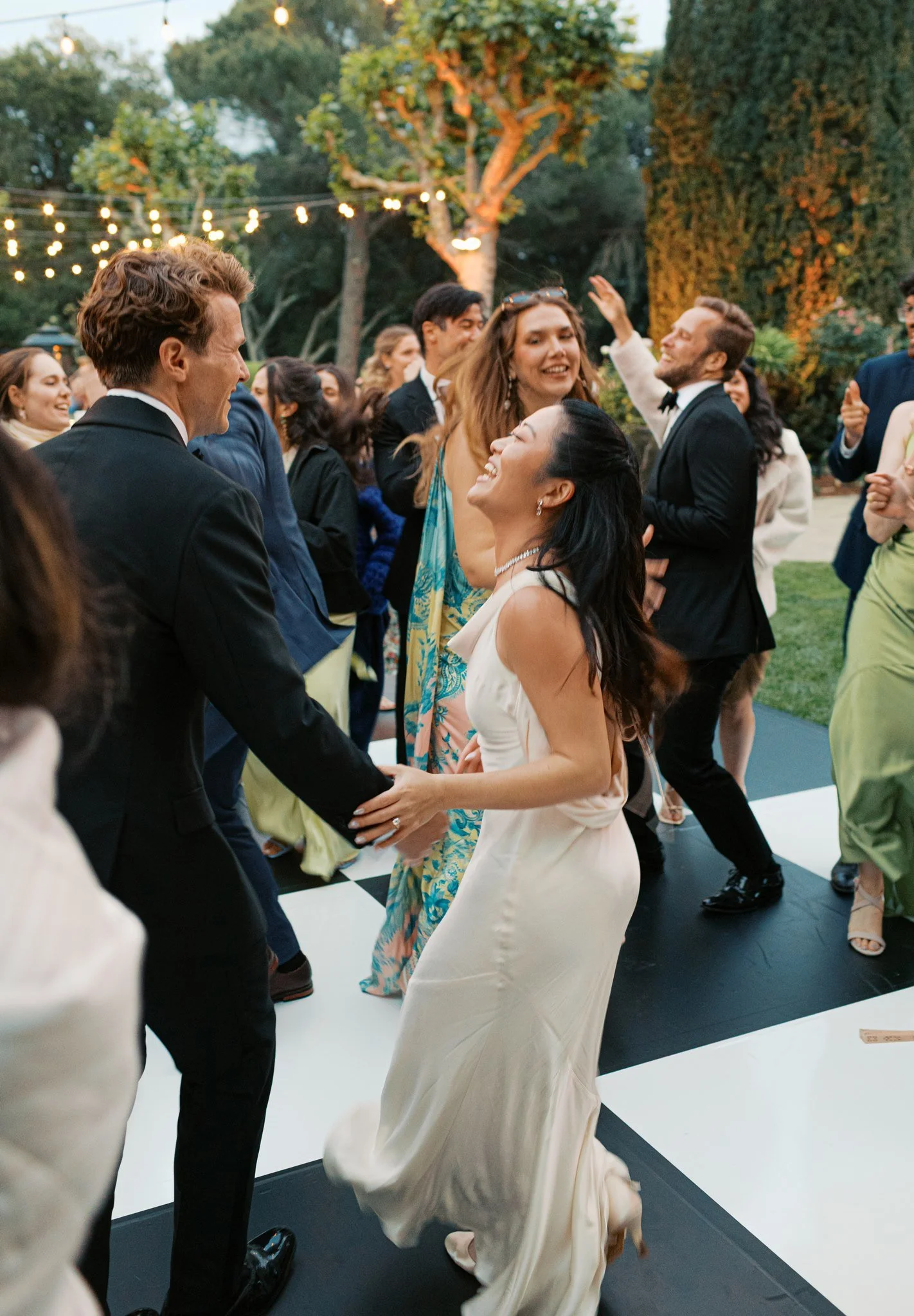People dancing at a party outdoors with string lights and trees in the background.
