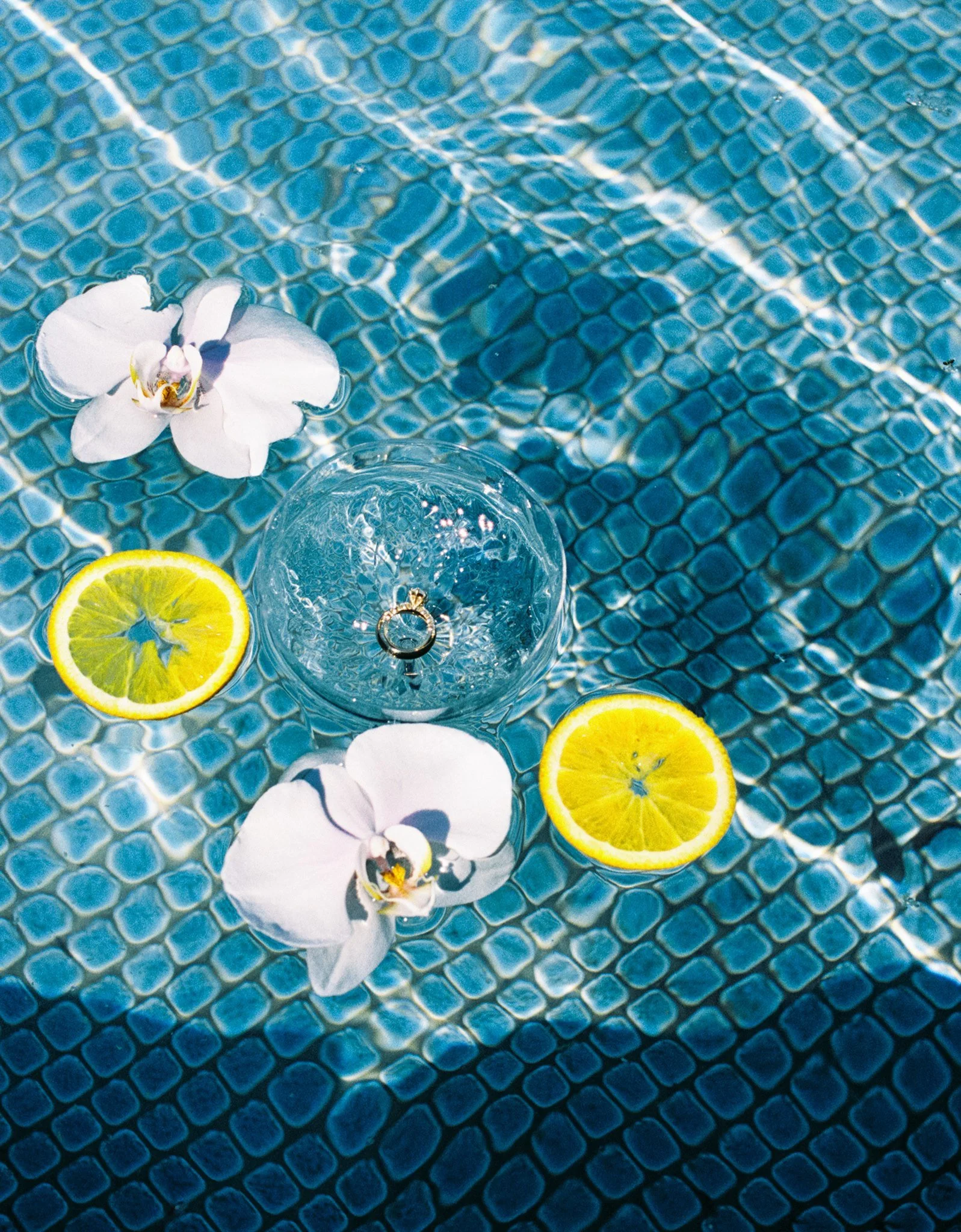 Close-up of a blue tiled surface with a white orchid flower, slices of lemon, and a glass of water with a ring in it.