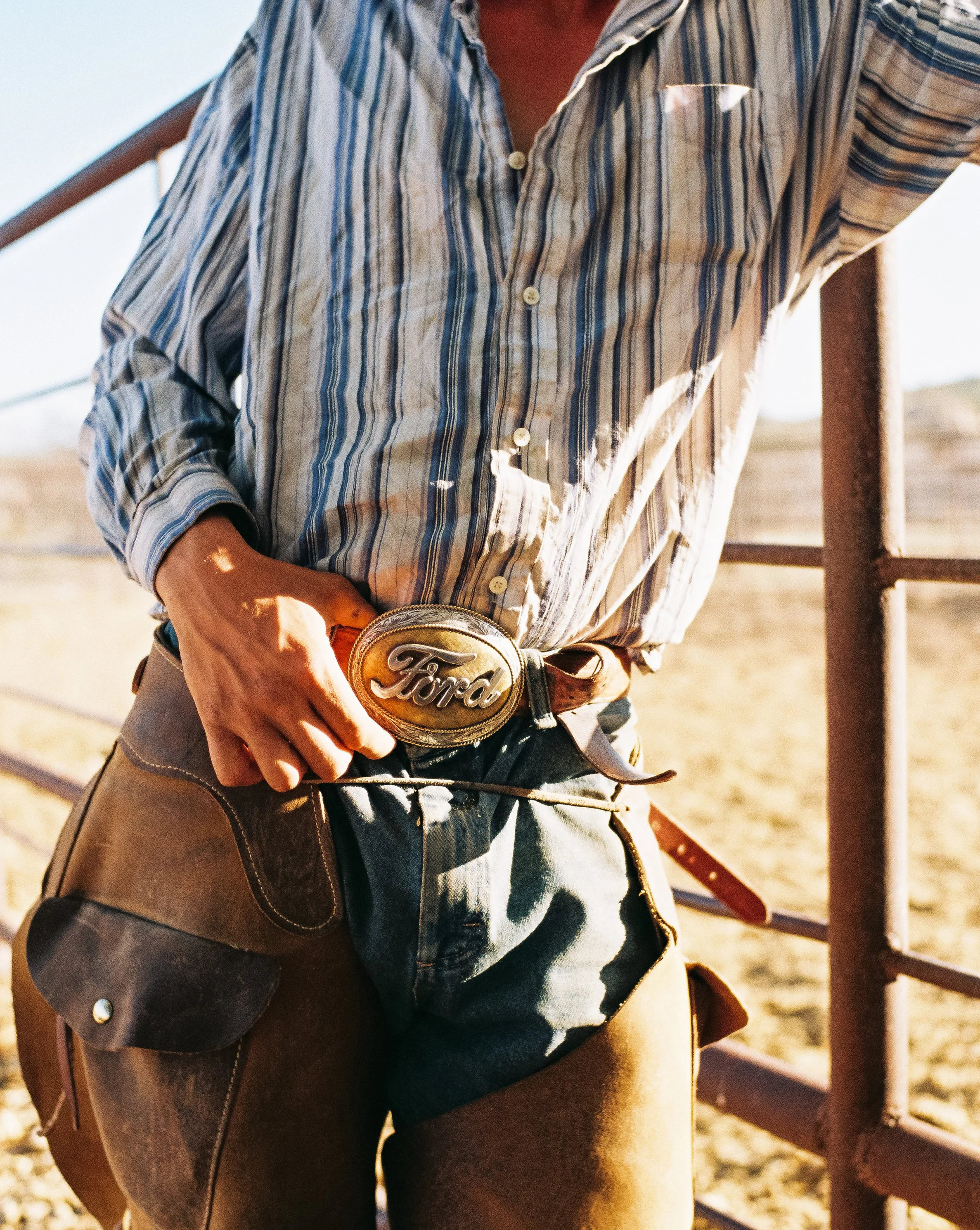 Man wearing a plaid shirt and leather chaps, adjusting a large Ford belt buckle in a rural, outdoor setting with a metal fence.
