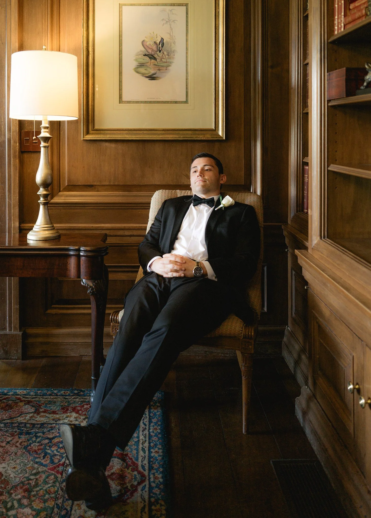 A man in a tuxedo with a white boutonniere sits relaxed with hands clasped in an elegant wood-paneled room, illuminated by a tall lamp and decorated with a framed artwork of flamingos and a palm tree on the wall.