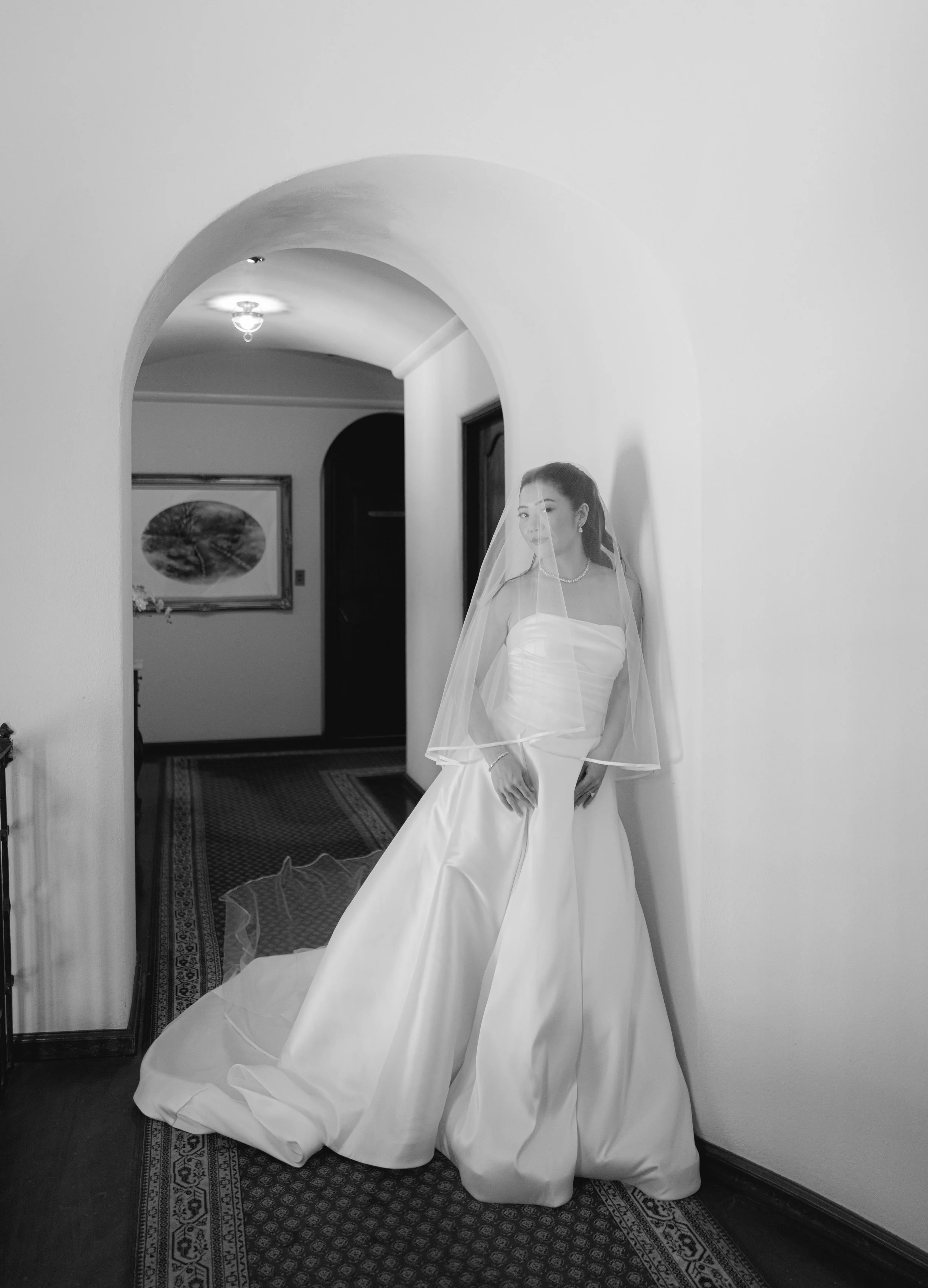 A bride in a wedding dress with a veil, standing in a hallway next to a white wall and a carpeted floor.