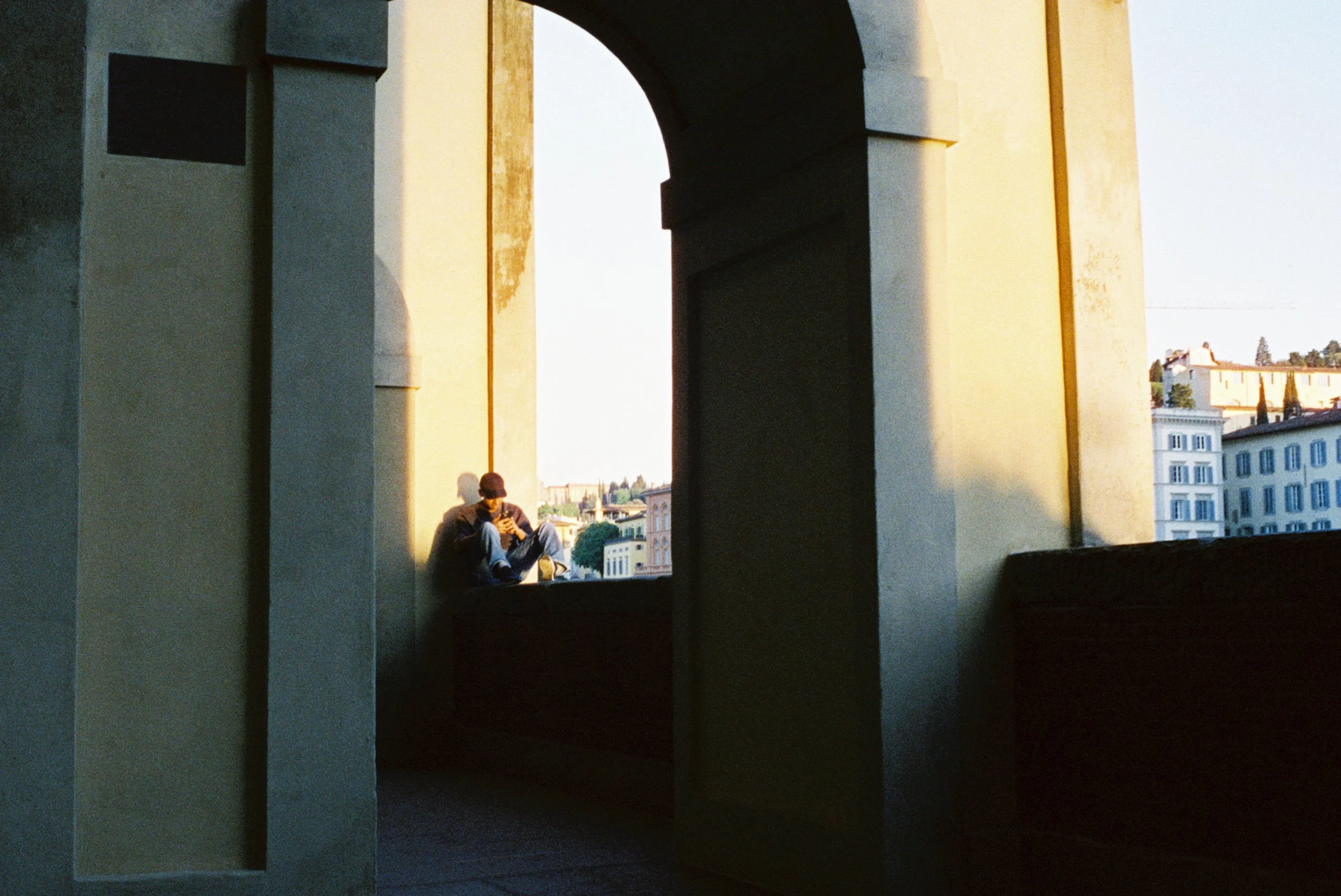 A person sitting on a ledge in an arched balcony, looking at a mobile phone, with a cityscape in the background during sunset.