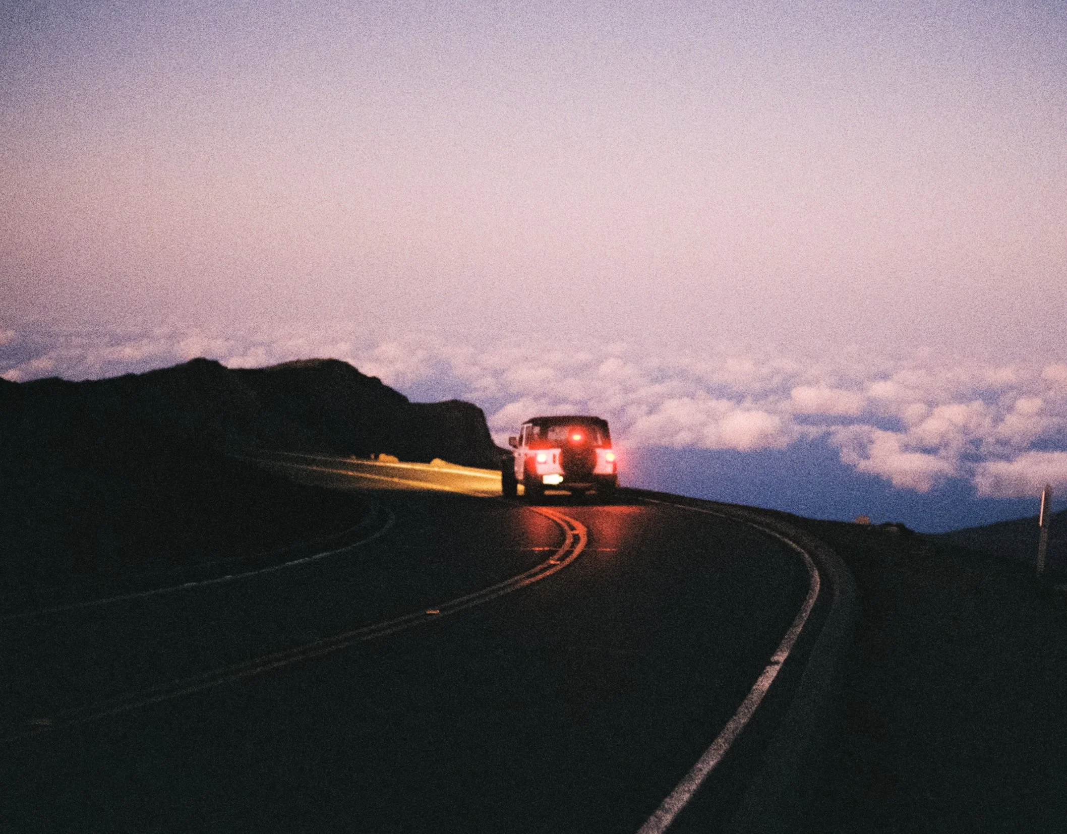 A car driving along a winding mountain road at dusk or dawn with a purple and pink sky and clouds in the background.