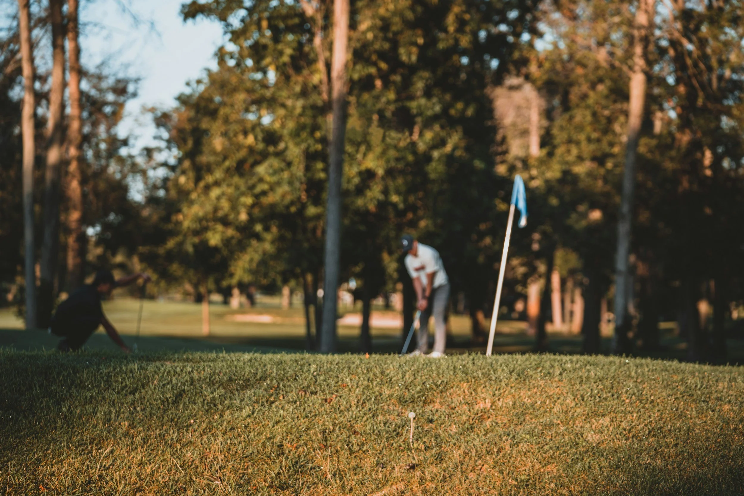 Two people playing golf on a green at a golf course during sunset, with trees in the background and a blue flag marking the hole.
