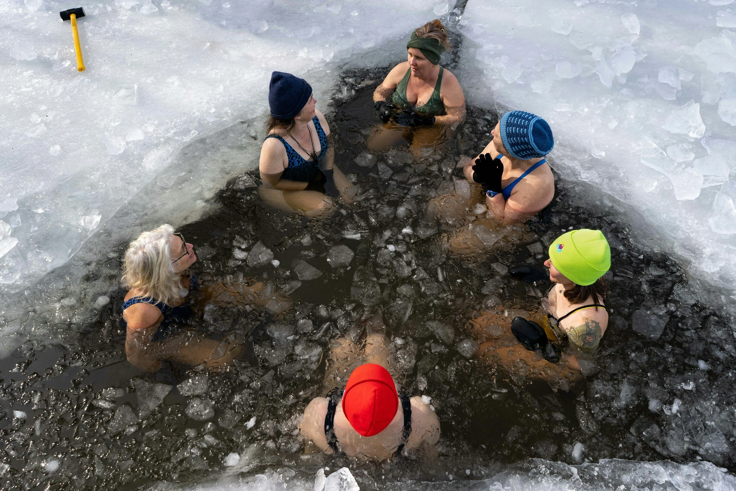 Seven women with colorful hats and gloves are standing in a small, ice-covered hole in a frozen body of water, engaging in a winter activity like ice swimming or bathing.