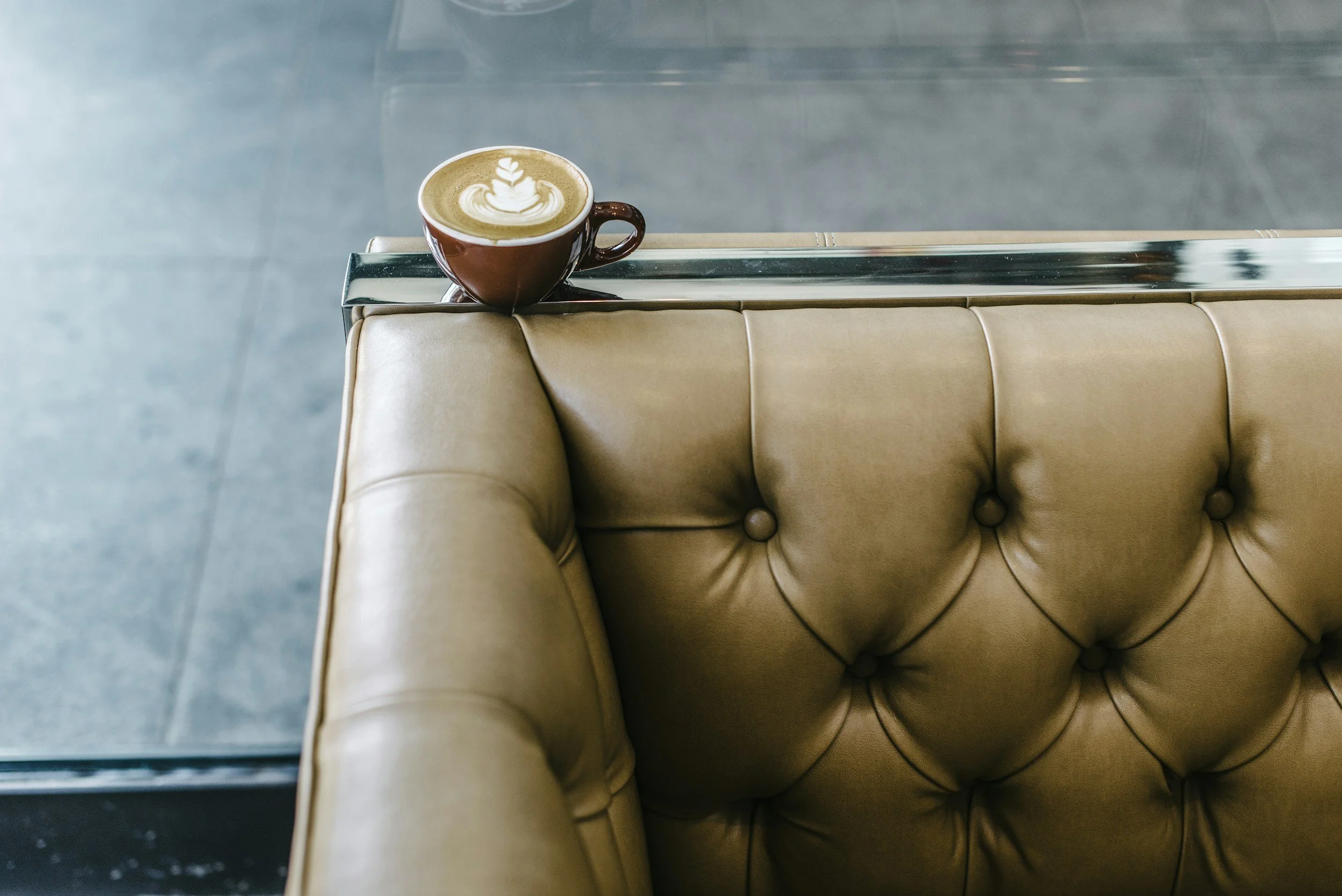 A cup of latte with latte art on a glass table next to a beige tufted leather sofa.