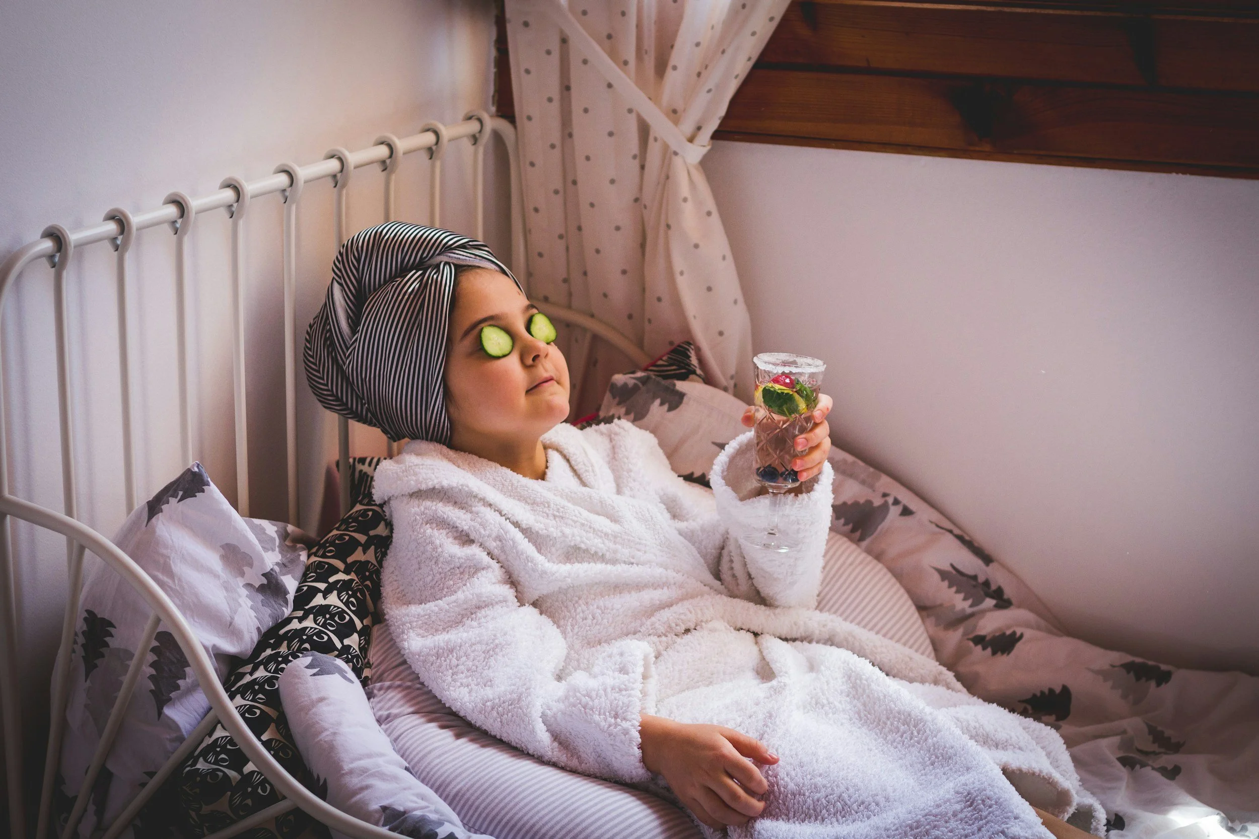 A young girl with cucumber slices on her eyes, wearing a striped headscarf, and a fluffy white bathrobe, sitting in bed and holding a glass of water with a slice of lemon and berries inside, in a cozy bedroom with pink bedding and polka dot curtains.