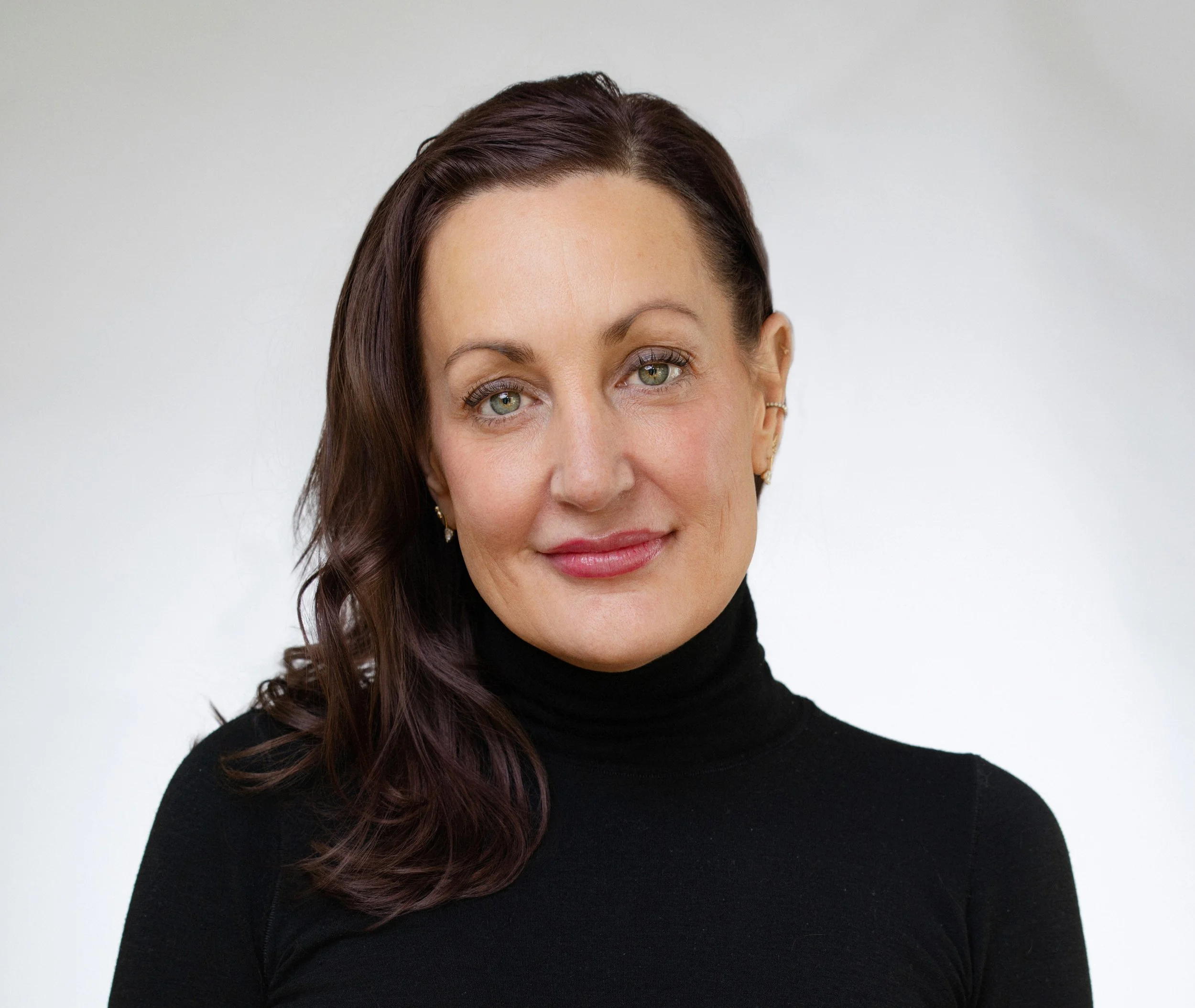 A woman with shoulder-length brown hair, wearing a black turtleneck, smiling against a plain light gray background.