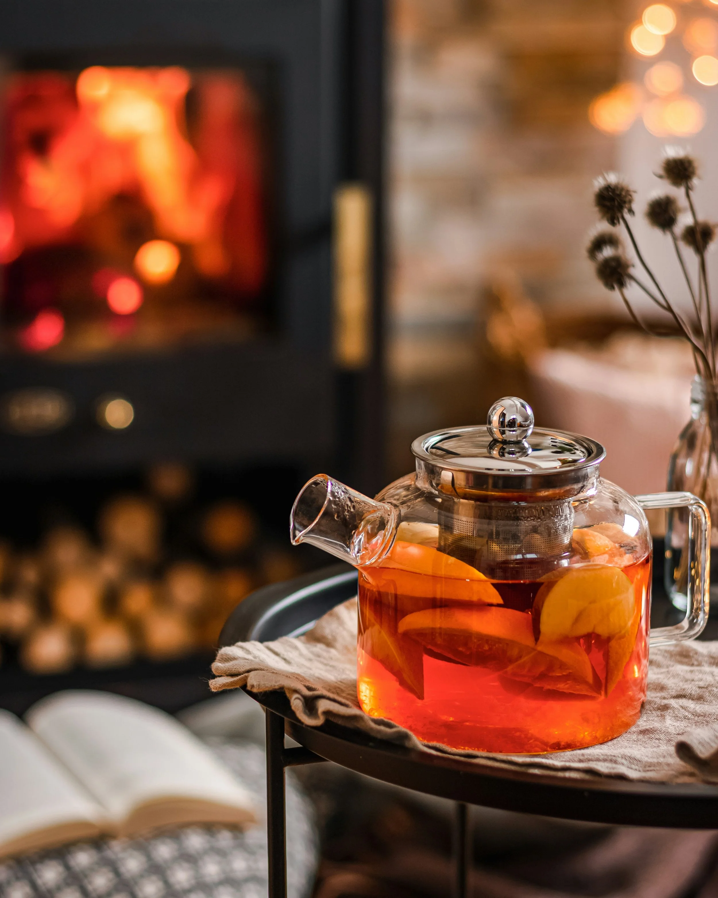A glass teapot filled with hot tea and lemon slices on a tray next to a book, with a fireplace in the background.