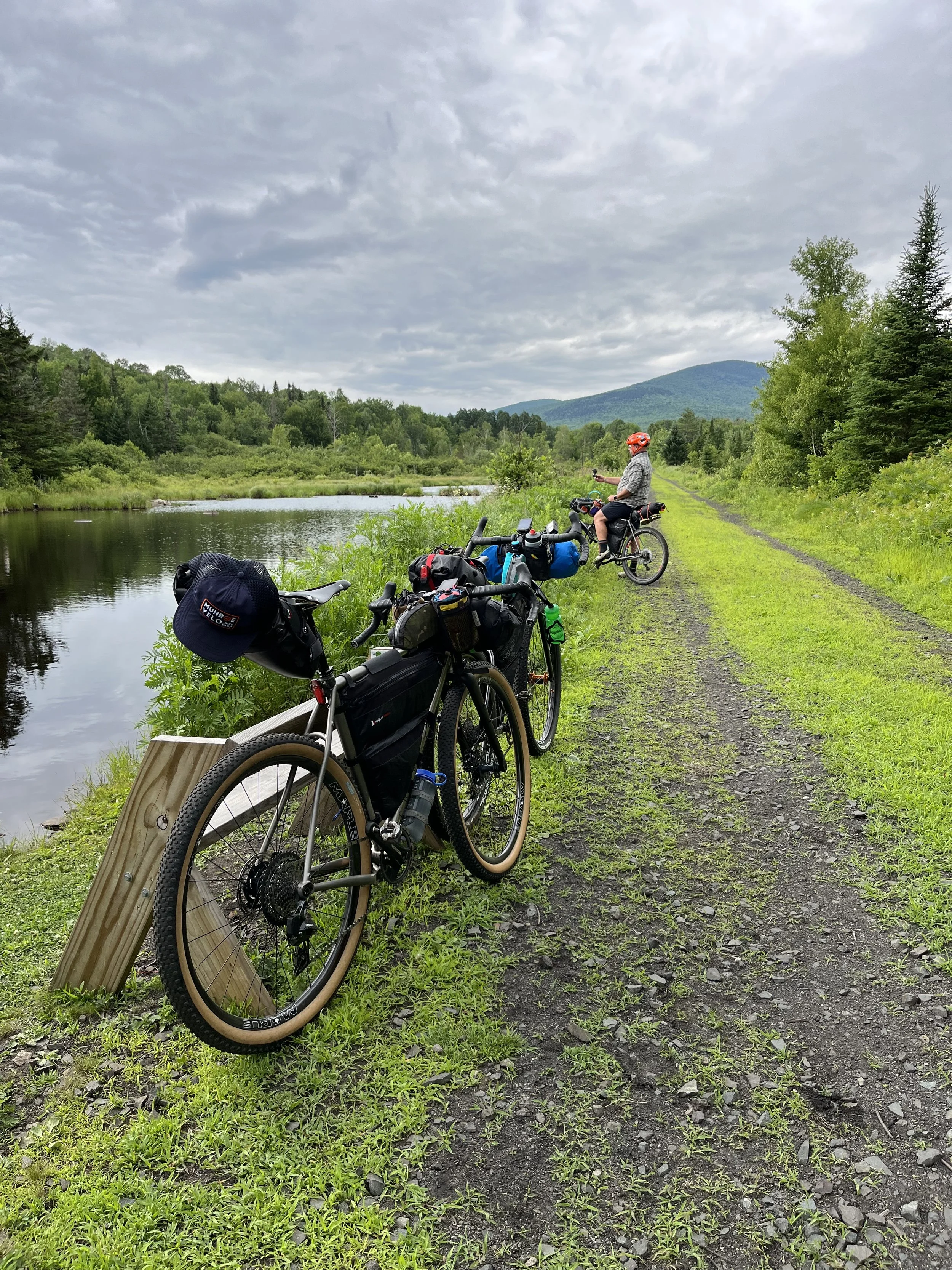 Two loaded bicycles parked along a green grassy trail beside a river, with a person wearing a helmet standing nearby, surrounded by lush trees and mountains in the background under a cloudy sky.