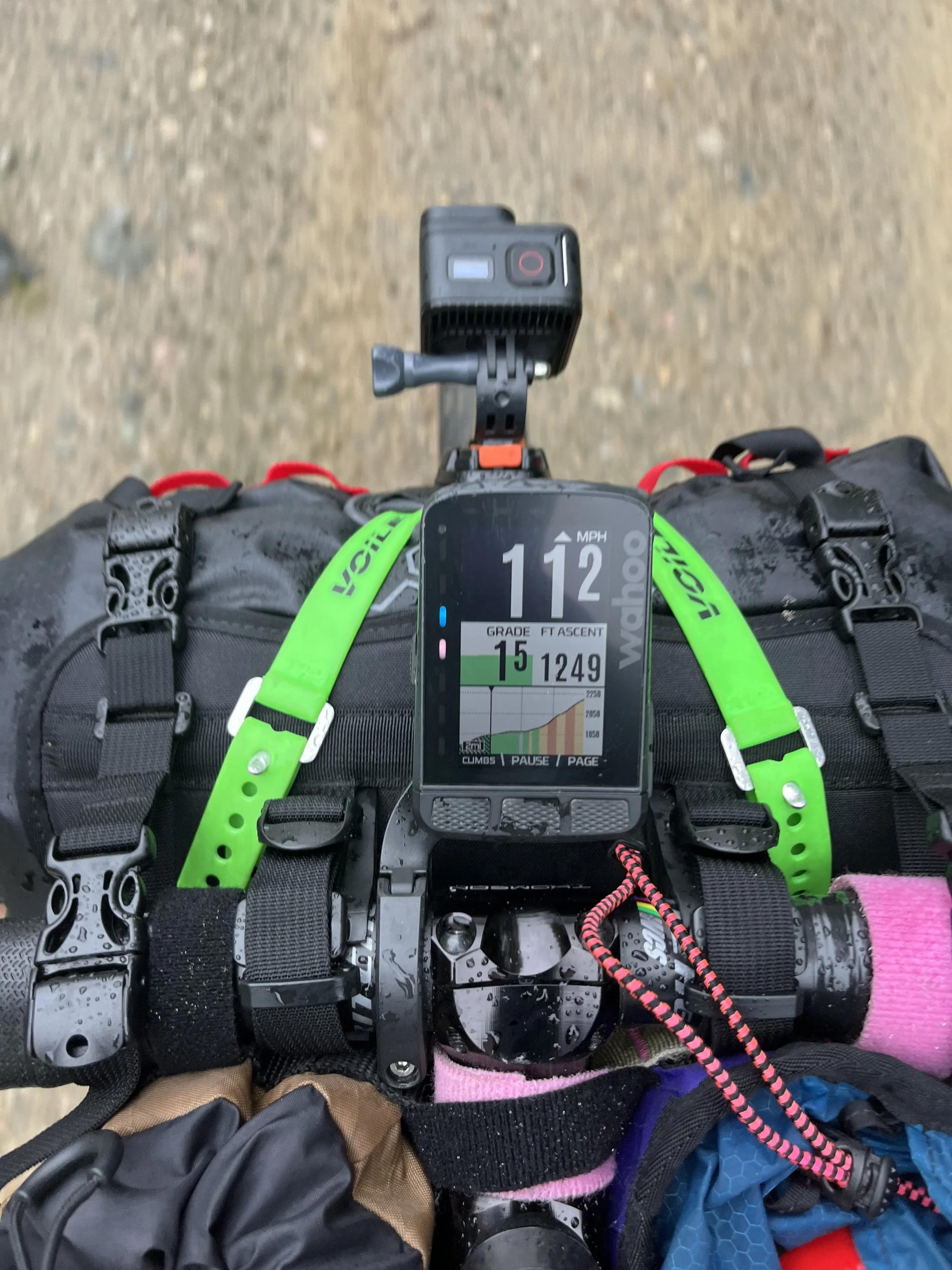 A bicycle handlebar with a mounted GPS device showing a speed of 112 miles per hour and an elevation of 1500 feet, rain droplets on the handlebar, and some gear and straps visible.