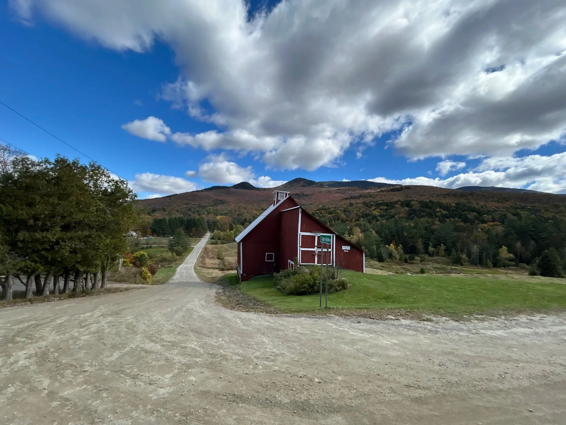 A rural scene with a gravel road leading past a red barn with white trim, surrounded by green grass and trees. Mountains with colorful fall foliage and a partly cloudy sky are in the background.