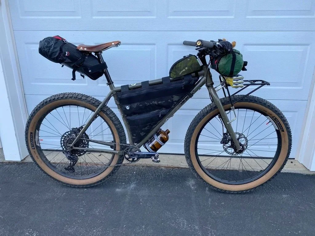 A mountain bike with tan tires, black frame, and gear bags attached, parked in front of a white garage door.