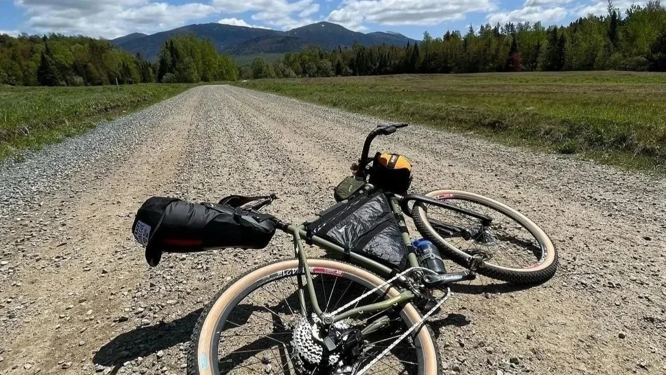 A mountain bike lying on a dirt road in a rural landscape with green fields, trees, and mountains in the background under a partly cloudy sky.