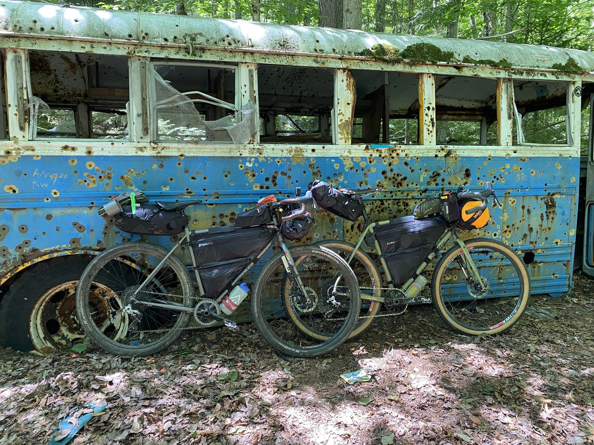 Two mountain bikes leaning against an old, rusted, and bullet-hole riddled blue bus in a wooded area, with fallen leaves on the ground.