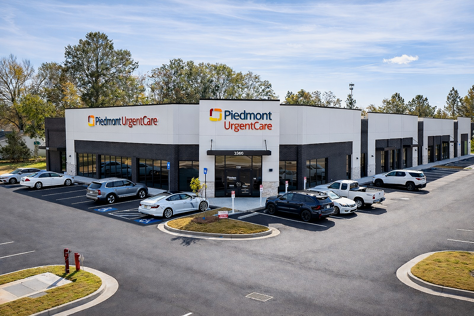 Exterior view of Piedmont Urgent Care medical facility with parking lot and several cars, trees in the background, and blue sky.