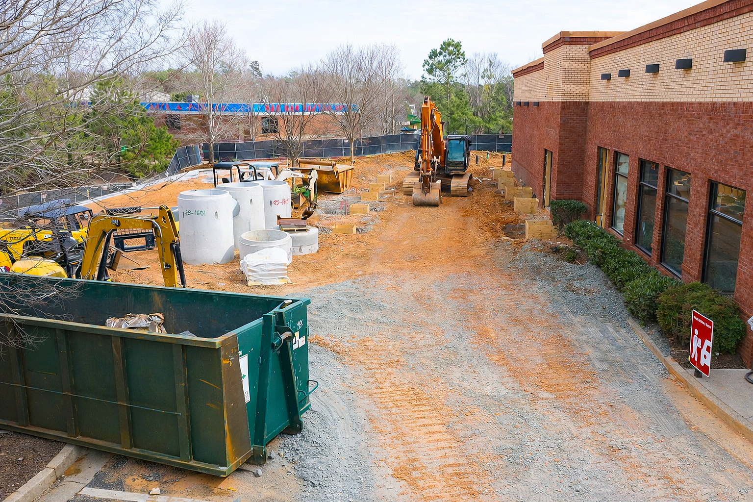 Construction site with an excavator, construction materials, and equipment next to a brick building, with a partially unpaved ground and trees in the background.